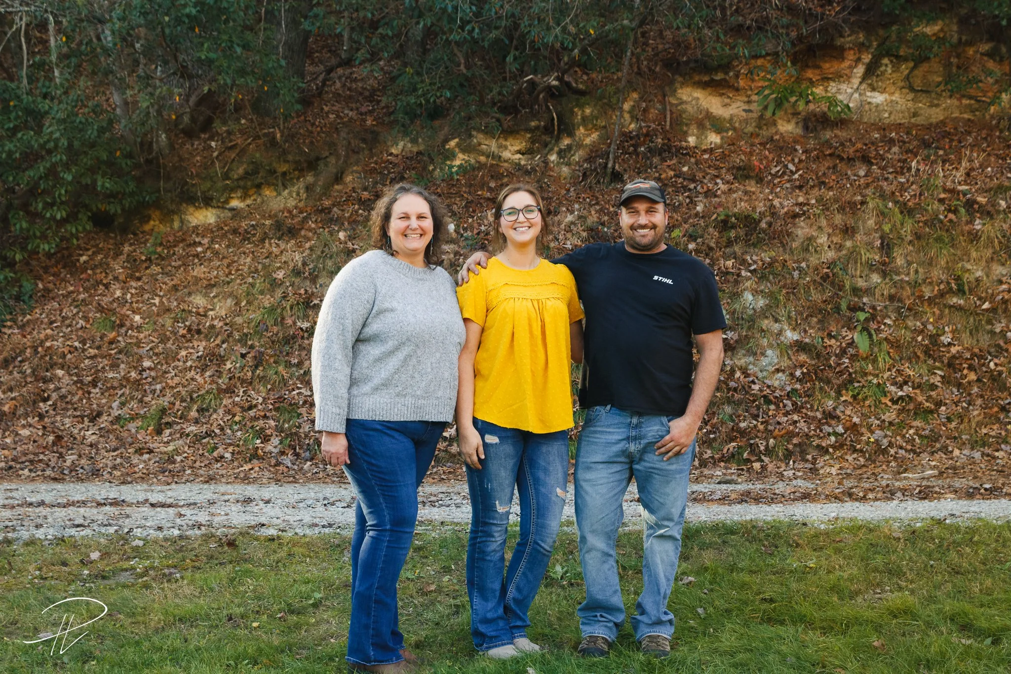 Three people standing outside on a grassy area with a wooded hillside in the background, smiling at the camera.