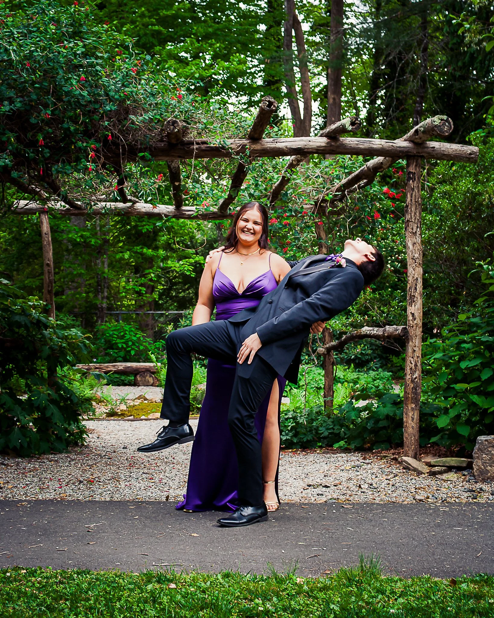 A young woman in a purple dress is standing on a gravel path, smiling and laughing, while a man in a black suit dips her backward in a dance move. They are outdoors with green foliage and trees in the background.