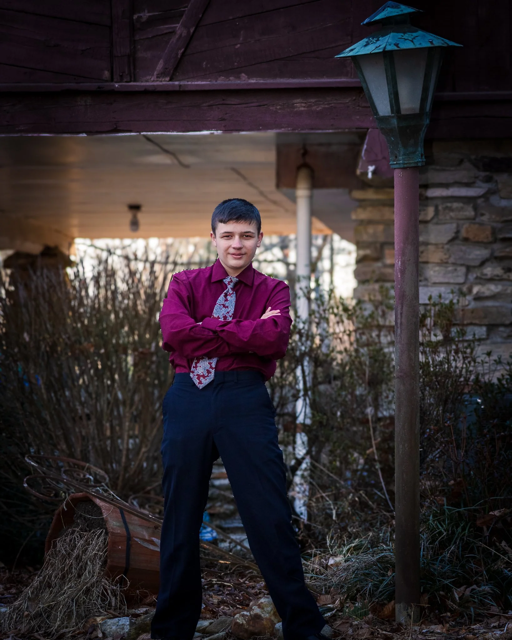 A young man with short dark hair wearing a maroon dress shirt, patterned tie, and dark pants, standing outdoors with arms crossed, in front of a rustic house with a stone wall and an old lamp post.