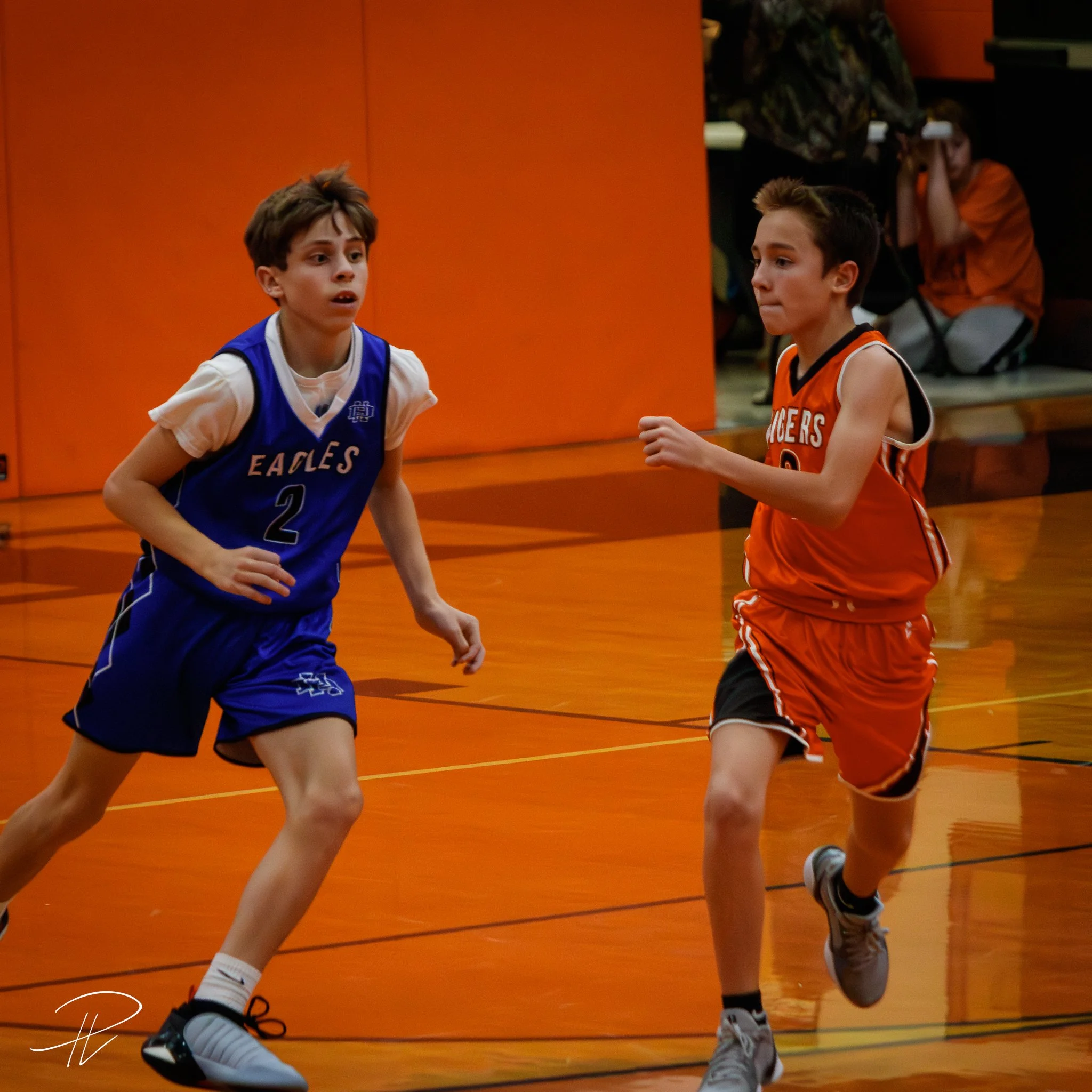 Two boys playing basketball on an indoor court, one in a blue jersey with 'EAGLES' and number 2, and the other in a red jersey, both running and focused on the game.