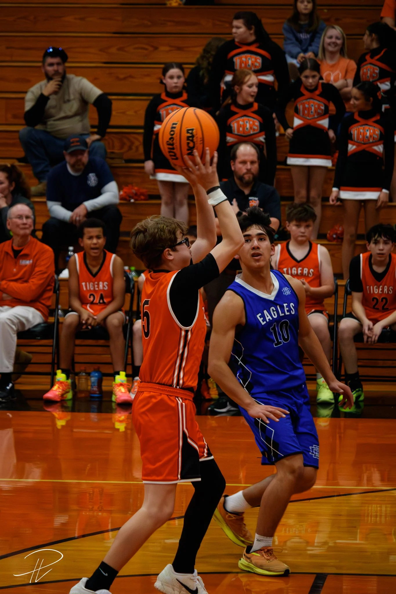 Young basketball player in orange jersey attempting a shot at the basket while a player in blue defends during a game. Spectators and cheerleaders are watching from the bleachers.