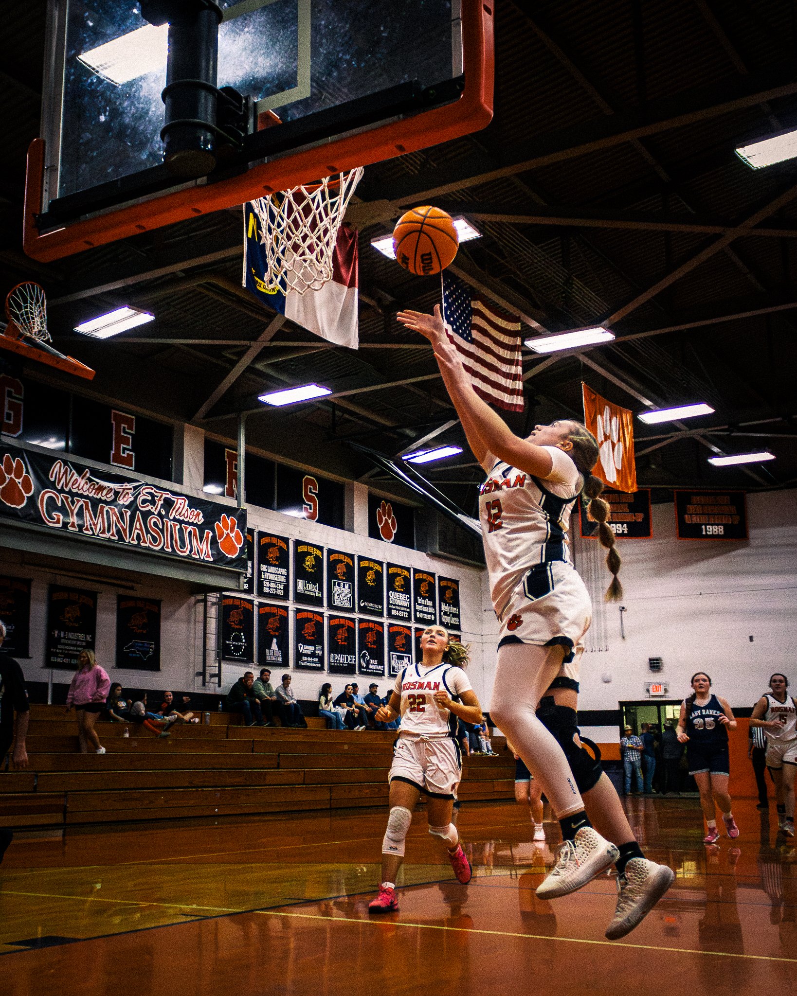 A female basketball player in a white and black uniform jumping to make a shot at the basket during a game at a gymnasium.