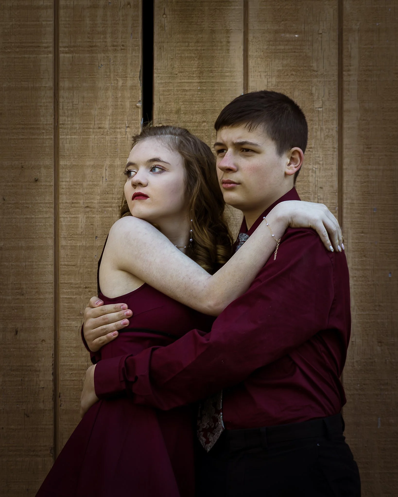 A young couple dressed in maroon clothing embracing against a wooden wall.