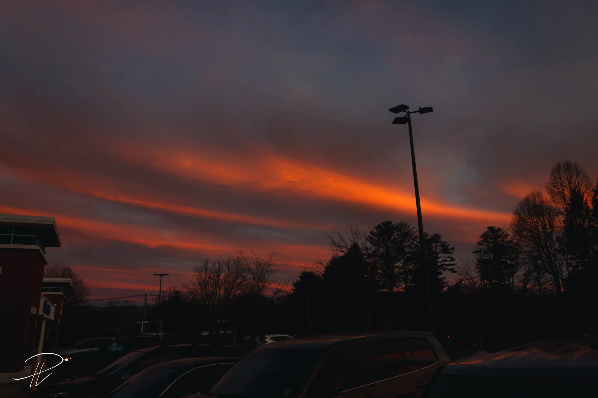 Sunset with clouds and orange, pink, and purple hues in the sky over a parking lot with cars, trees, and a lamppost.