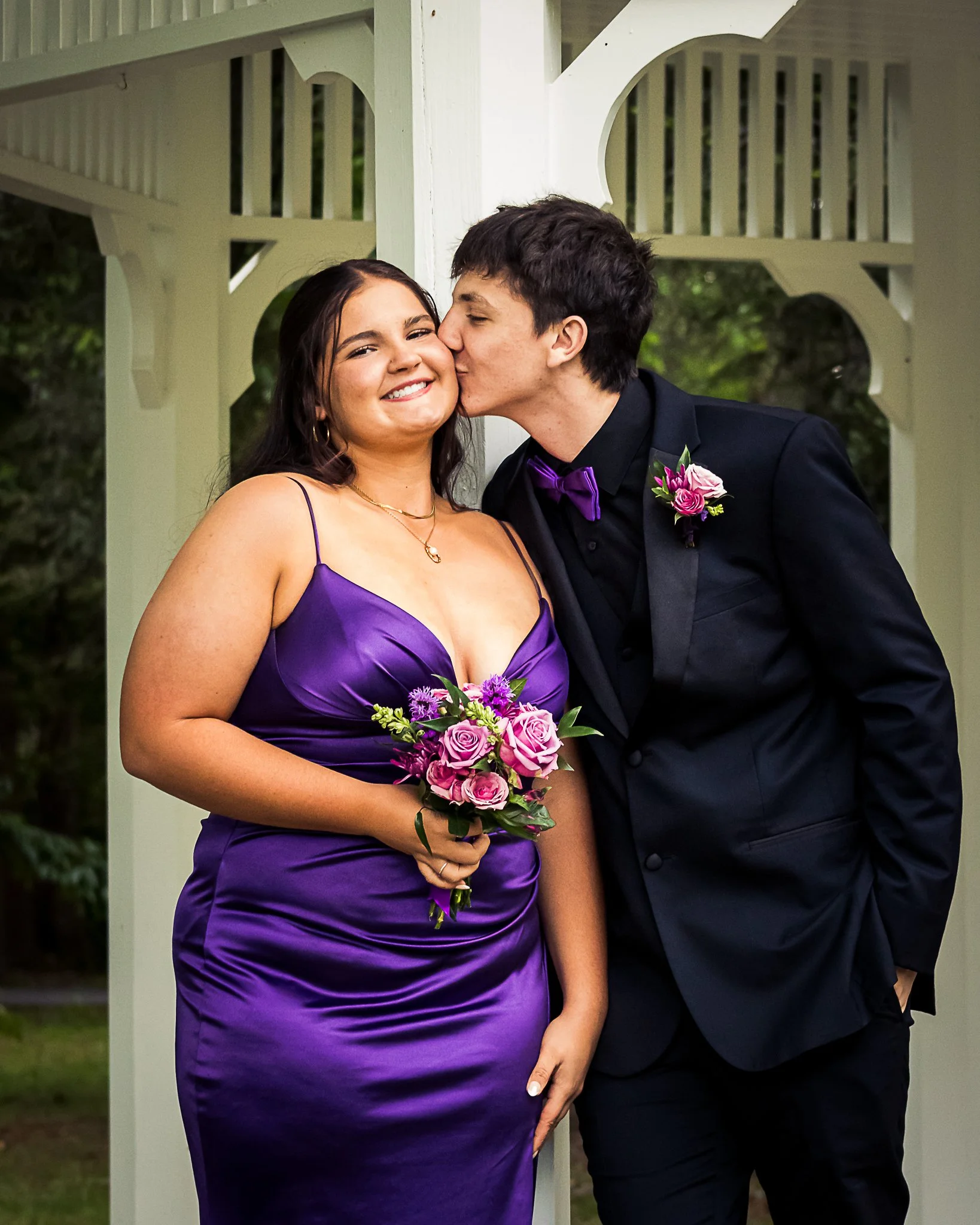 A young couple at a wedding, with the woman in a purple dress holding a bouquet and the man in a black suit with a purple bow tie, sharing a kiss on the cheek.