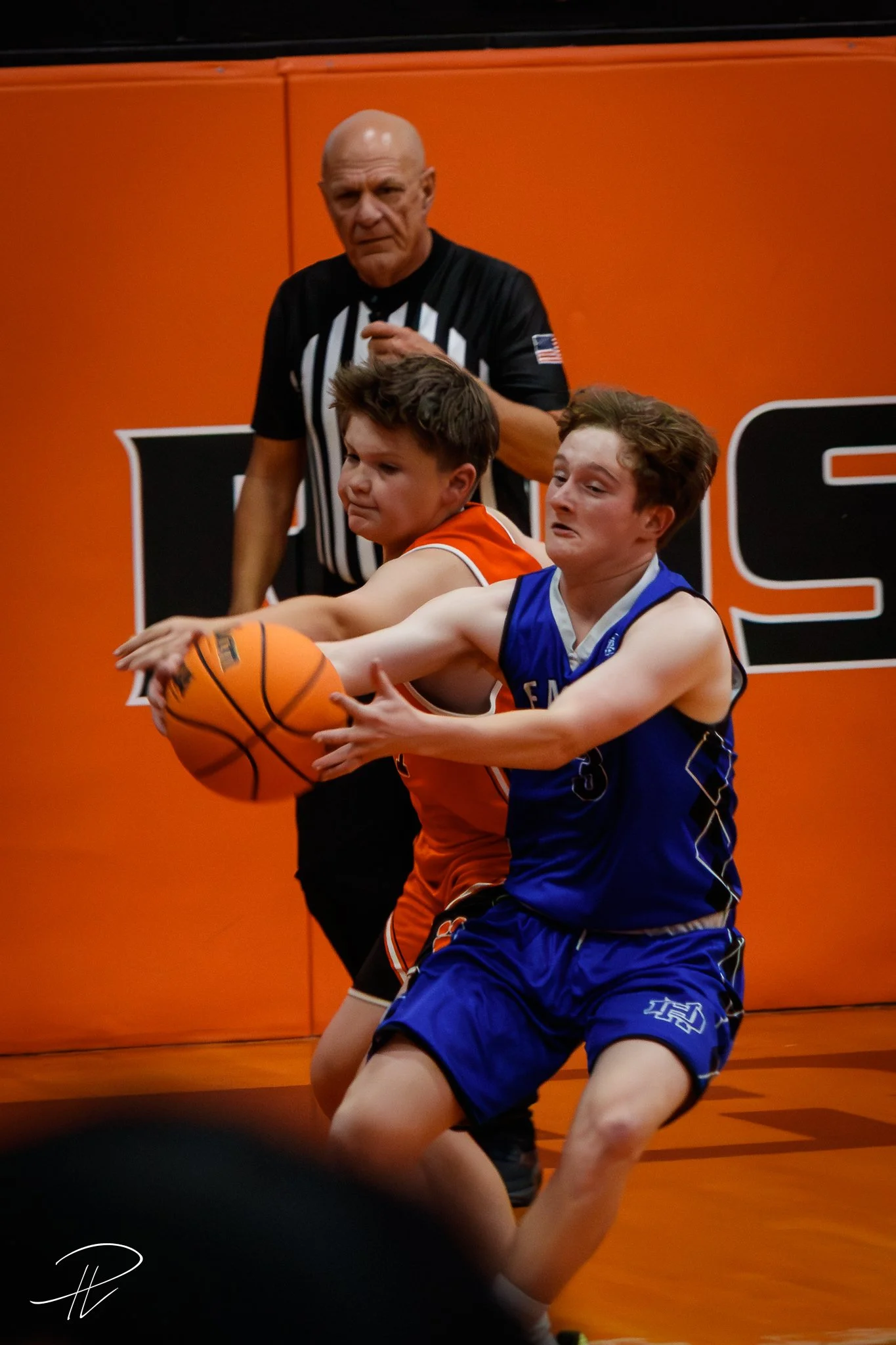 Two boys playing basketball, one in an orange jersey and the other in a blue jersey, fighting for possession of the basketball with a referee watching in the background.