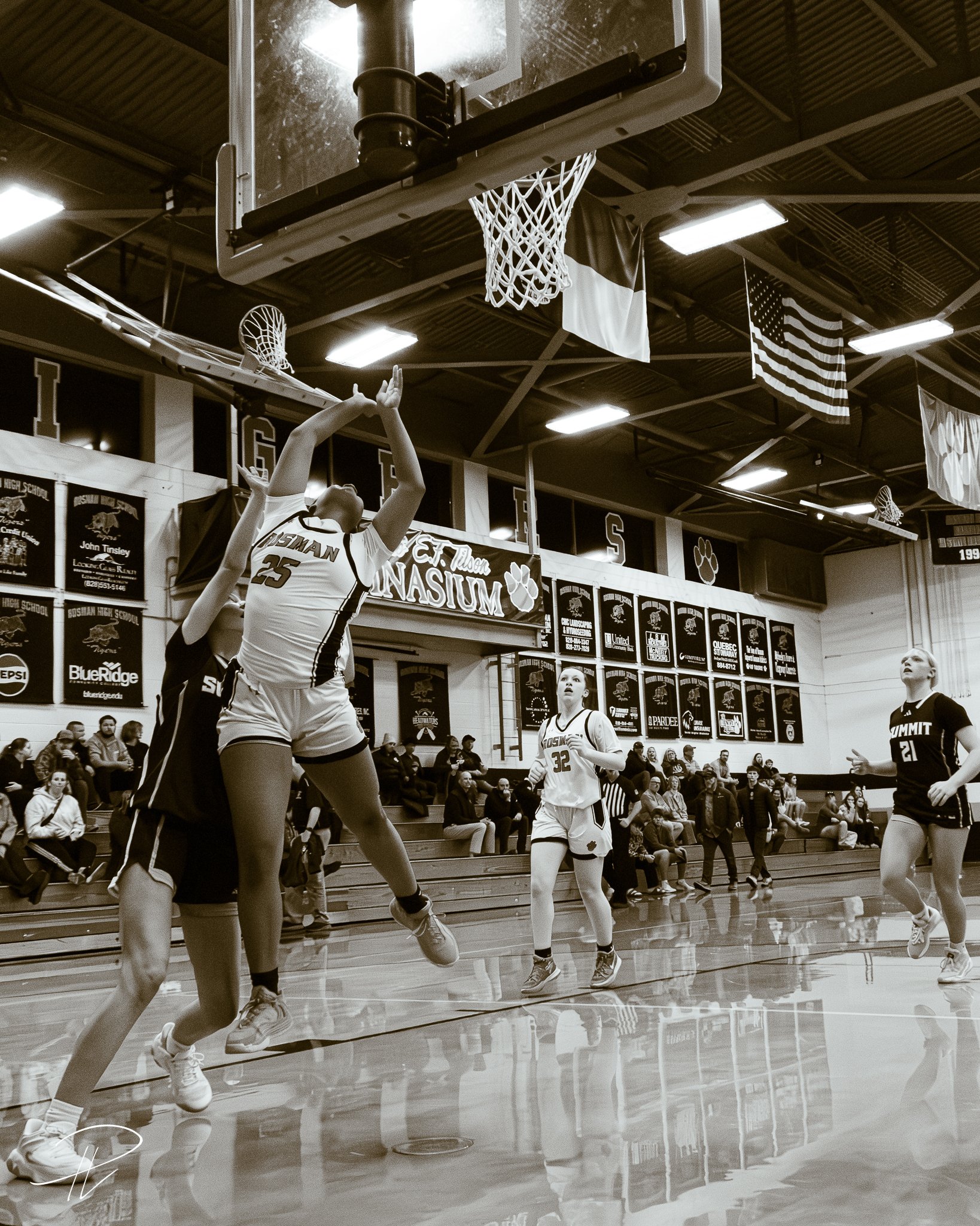 A girls' basketball game in a gymnasium with players in action, one jumping to shoot or block near the basket, when the gym is filled with spectators and banners hanging on the walls.