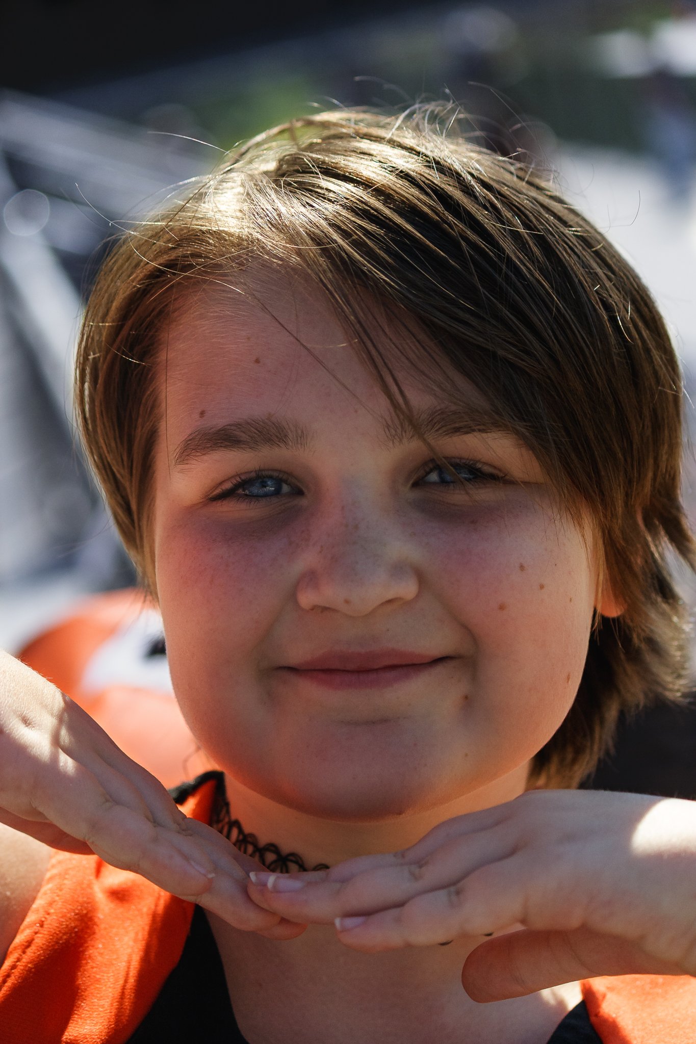 A young person with short brown hair, blue eyes, and freckles on their face smiling and holding their hands under their chin, outdoors in bright sunlight.