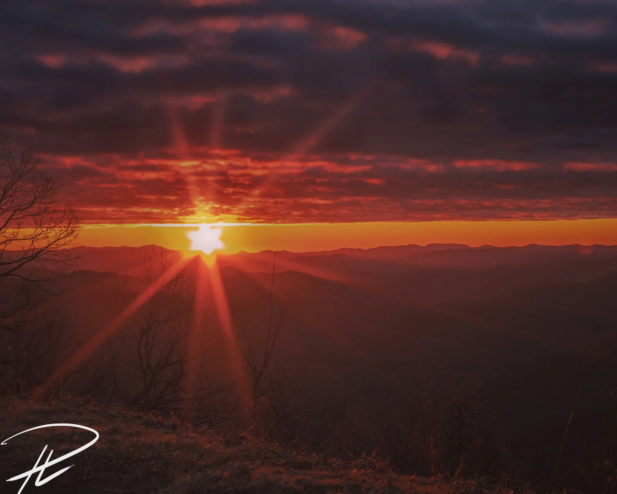A sunset over a mountain landscape with dark clouds and rays of sunlight breaking through, casting an orange and red glow across the sky.