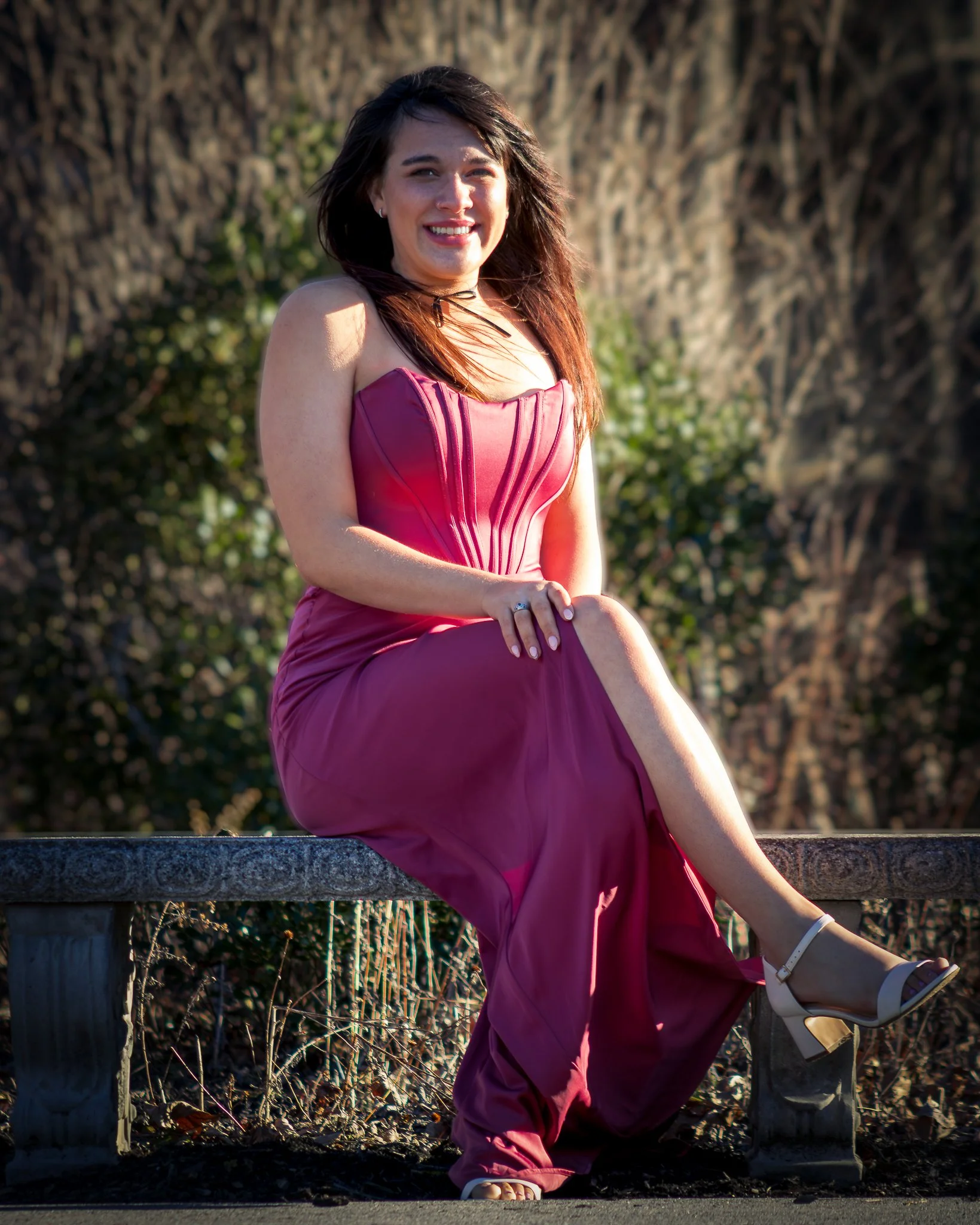 A woman in a pink dress sitting on a stone bench outdoors, smiling at the camera with a background of trees and bushes.