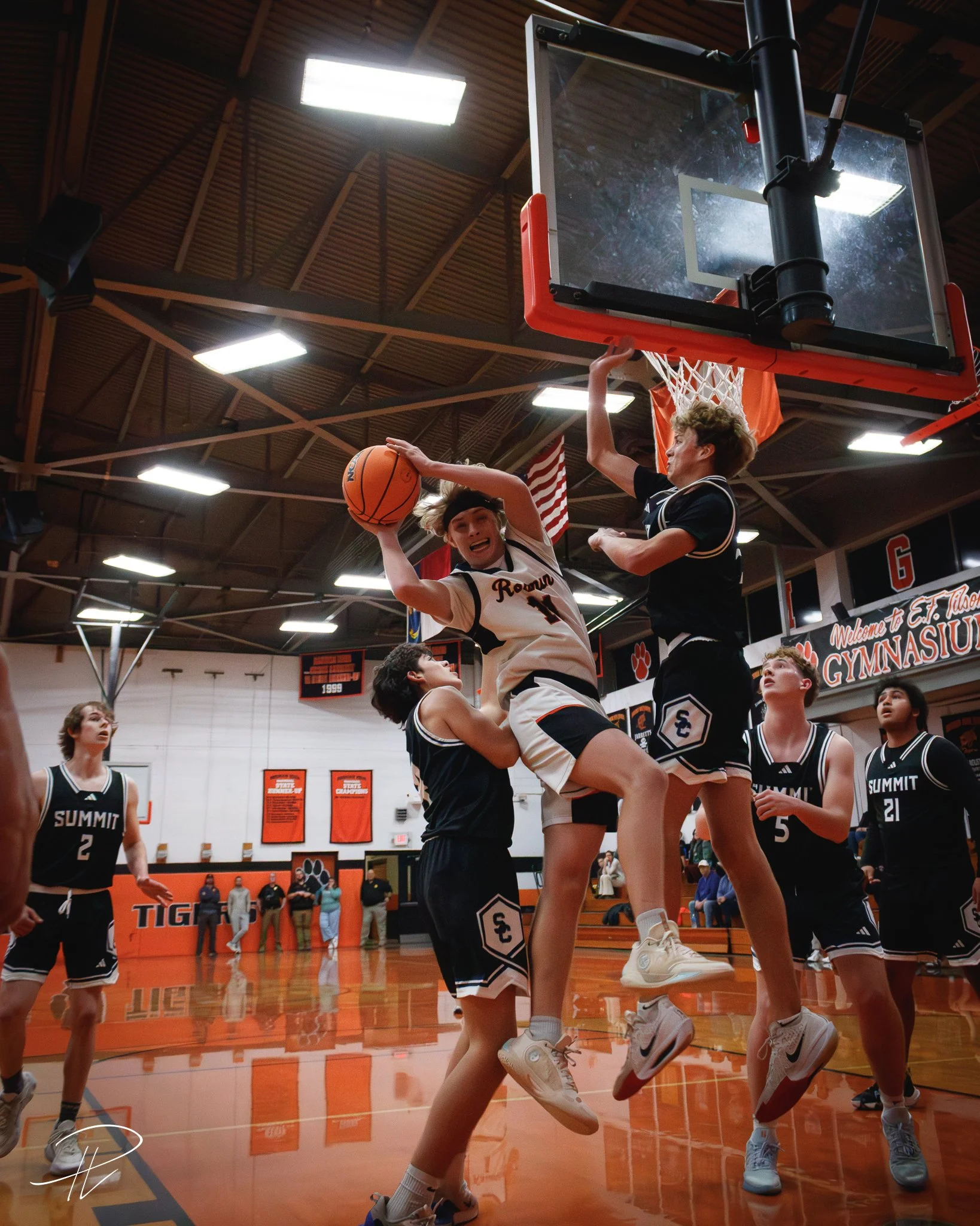Three basketball players jumping near the hoop; one in white jersey with number 11 holding the ball, two in dark jerseys attempting to block.