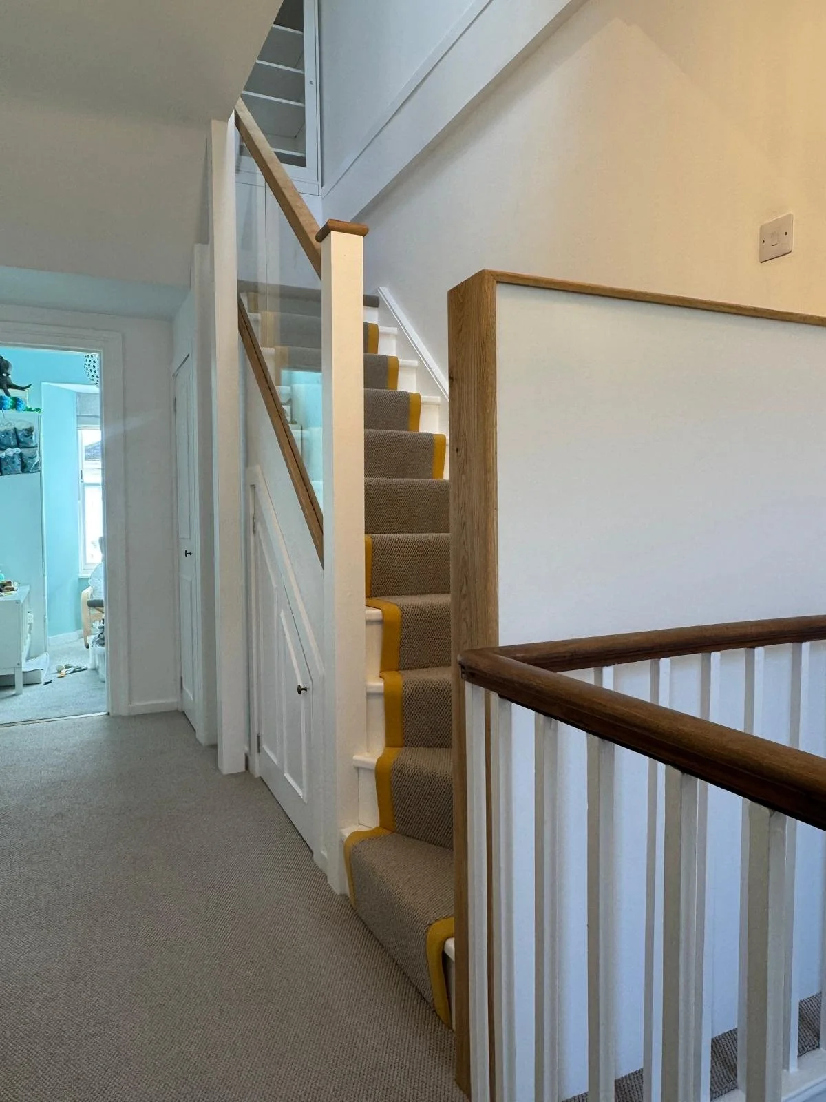 Interior view of a staircase and hallway in a house, with beige carpeted stairs, white walls, and a wooden handrail.