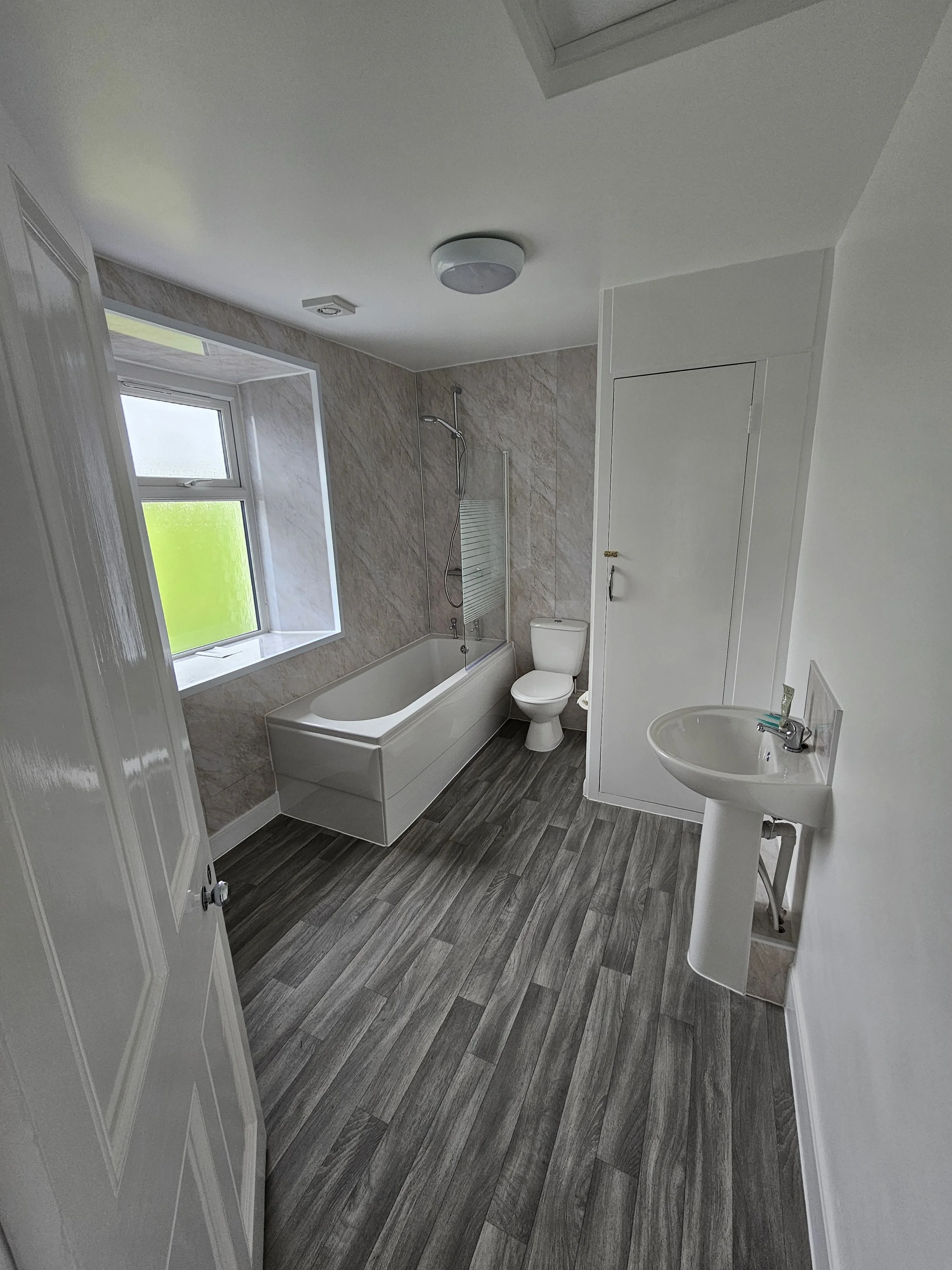 A bathroom with grey wood-look flooring, a white bathtub with a mounted showerhead, a white toilet, a small white pedestal sink, a window with frosted glass, and a ceiling light fixture.