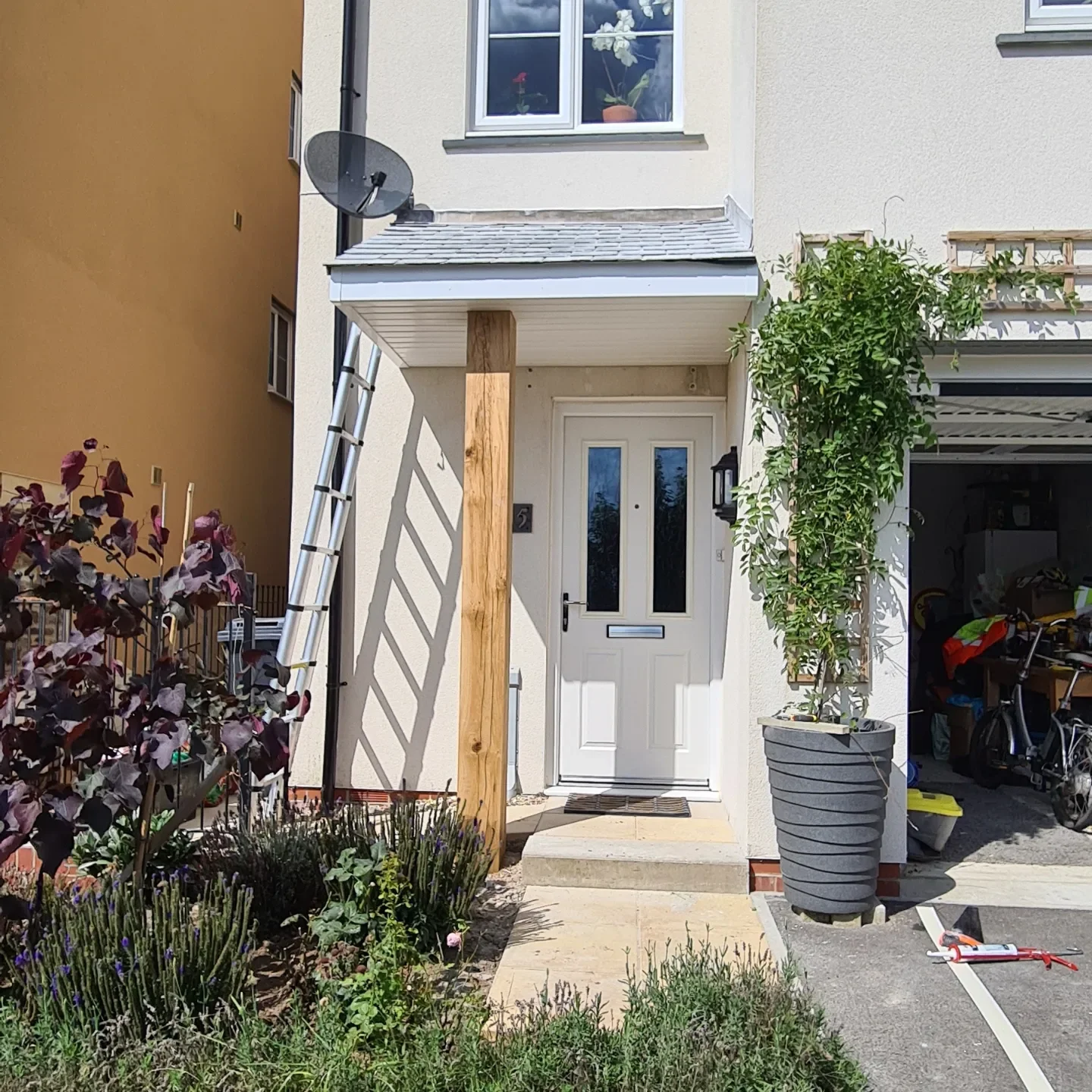 The front entrance of a house with a white door, a small porch, and a garden with various plants. There is a ladder leaning against the wall, a satellite dish above, and a large potted tree on the right side. To the right, a garage is visible with bi