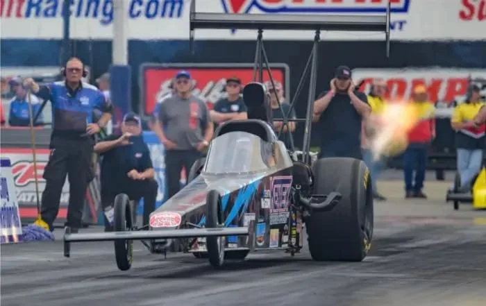 Top fuel dragster car on a race track with a group of people in the background, some wearing racing uniforms and others in casual clothing, and a fiery exhaust flame coming from the rear.