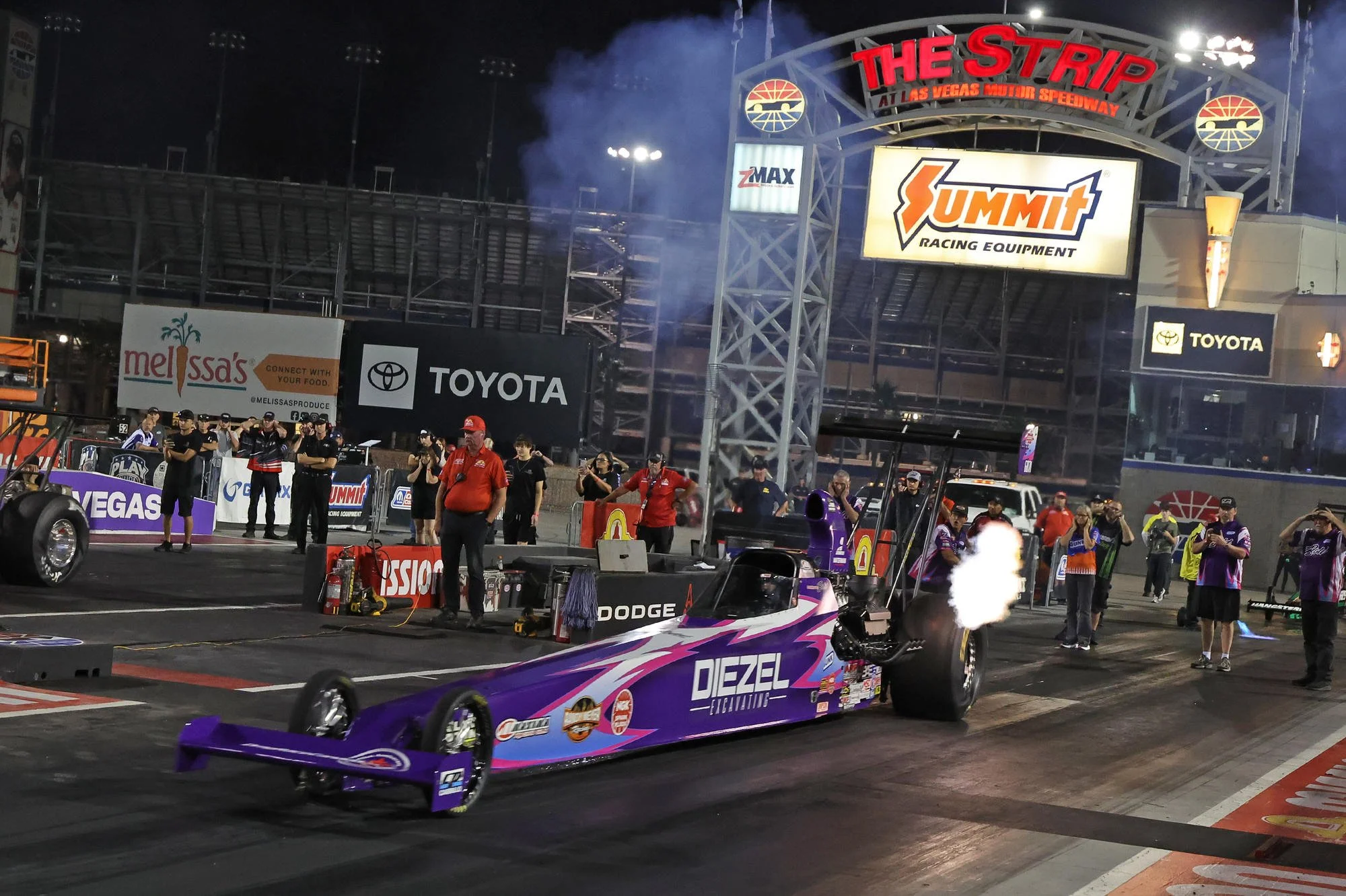 A drag racing car with a purple and pink paint job on the starting line at Las Vegas Motor Speedway, with team members and spectators in the background, and signs for Summit Racing Equipment and Toyota.