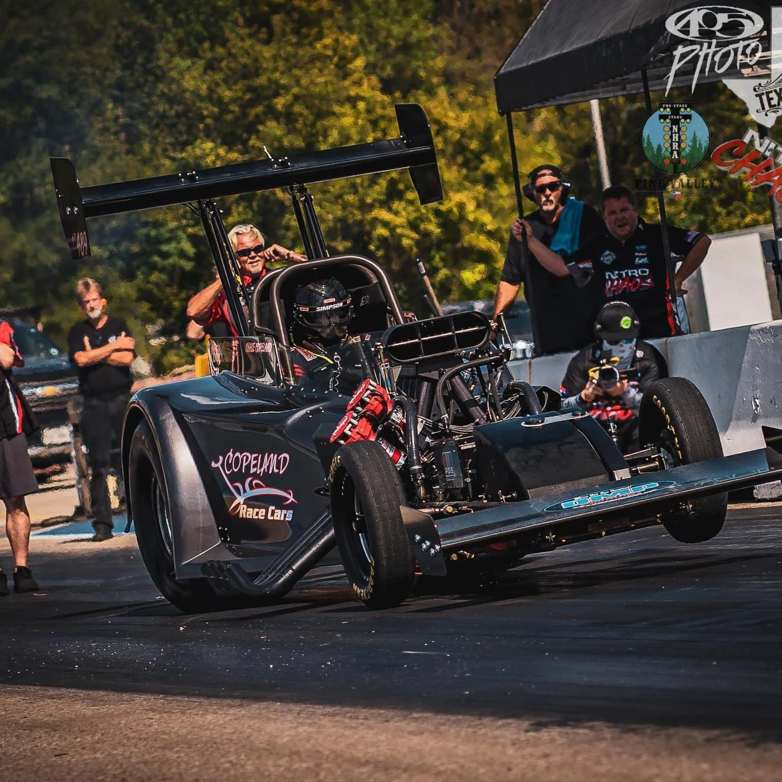 A black race car with a large rear wing, on a drag strip, surrounded by team members and officials.
