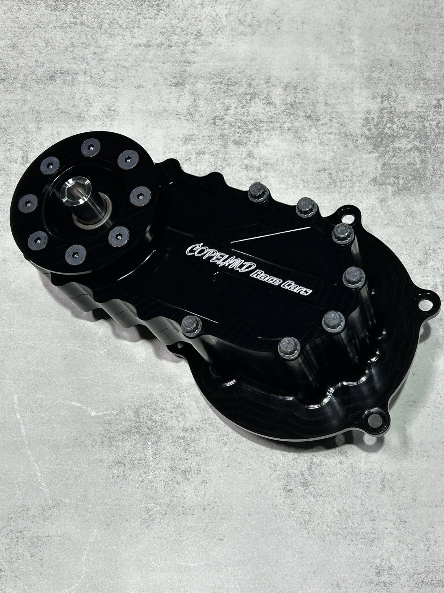 Black automotive part with bolts, labeled 'COPLELAND Race Cars,' on a gray surface.