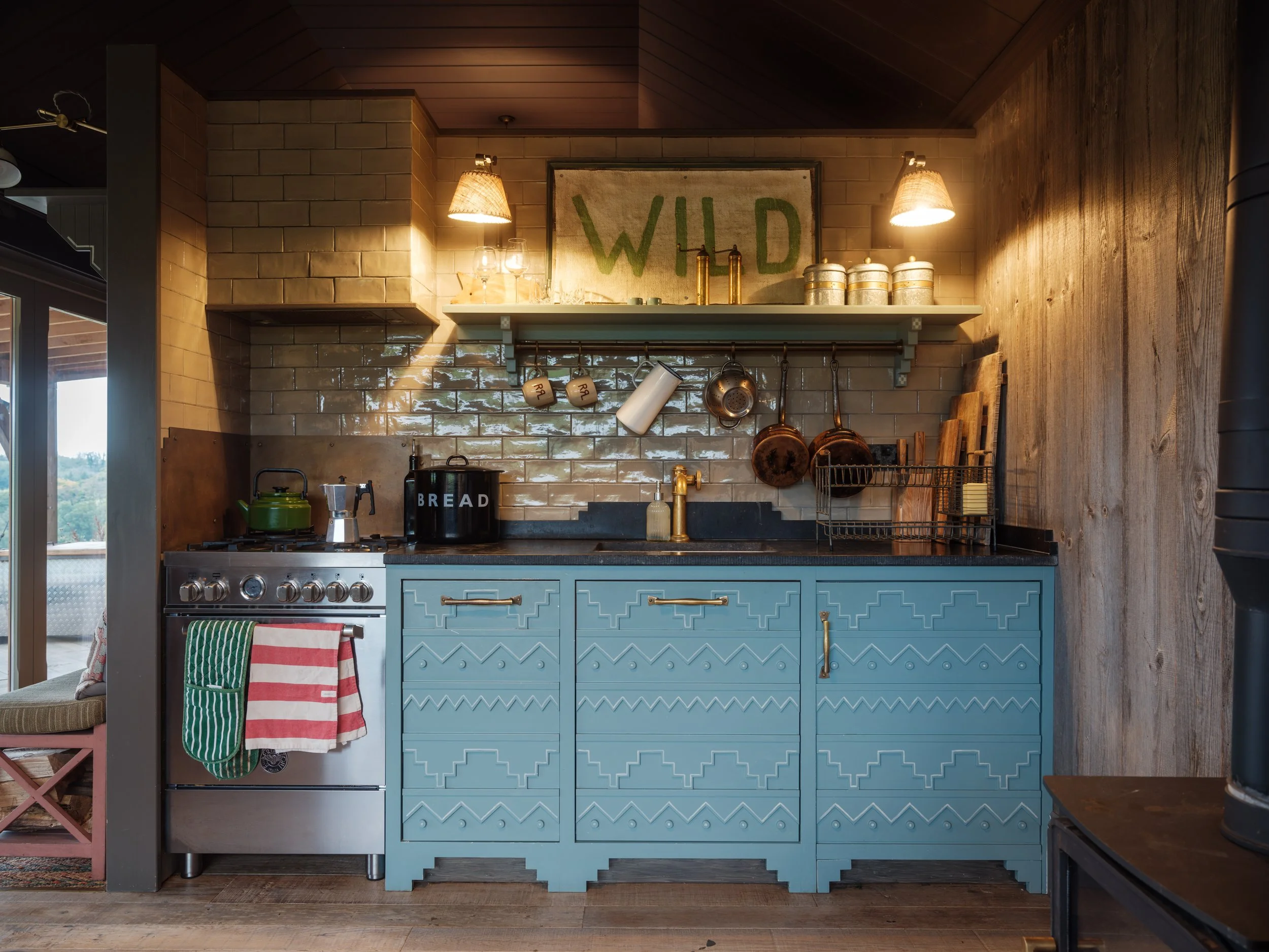 A rustic kitchen with blue cabinets, a black countertop, and a beige tiled backsplash. There is a stove with a green kettle, a bread box, and various hanging pots and mugs. A sign reading 'WILD' is on the wall above the shelf. The lighting is warm an