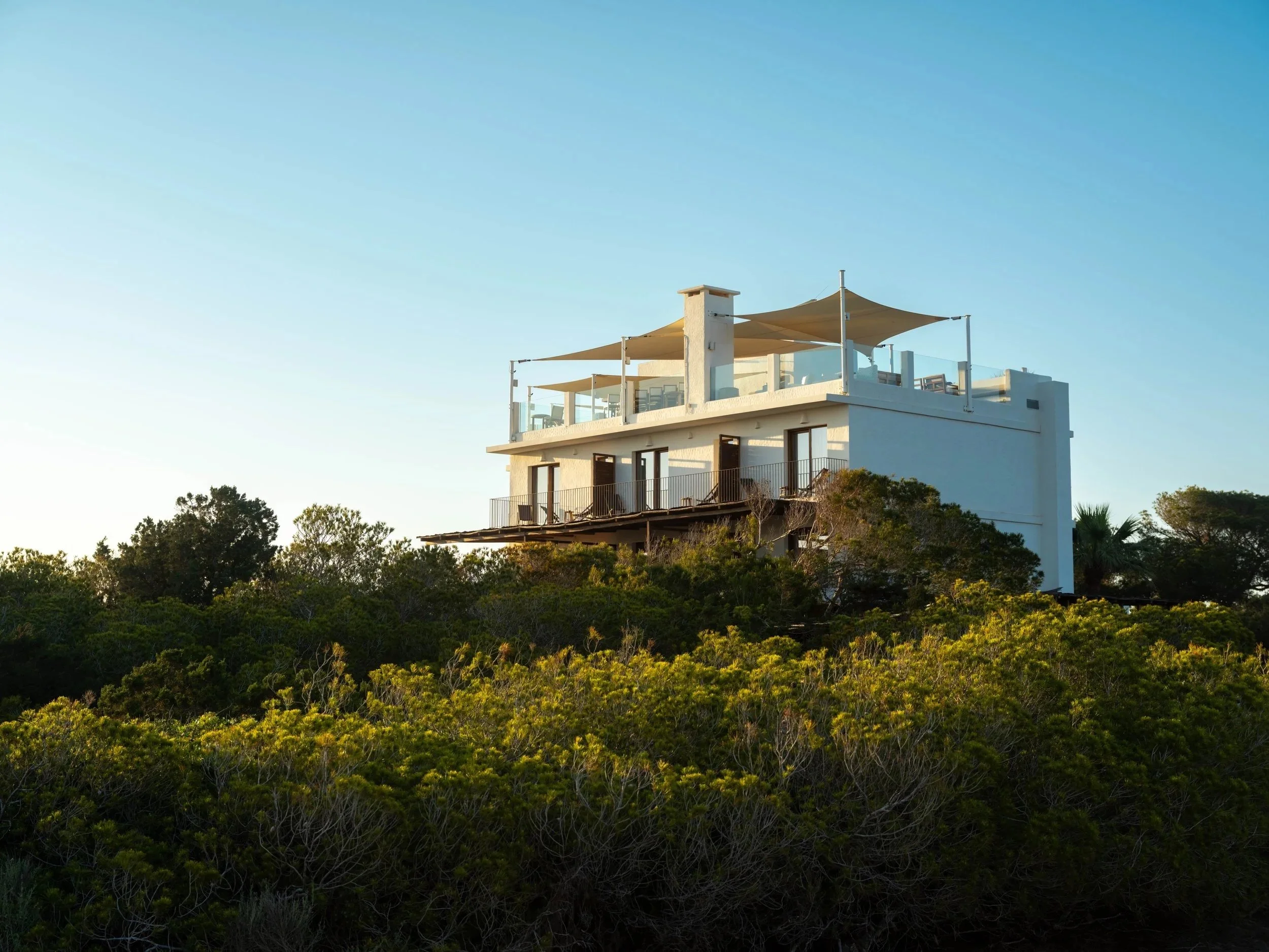 White multi-story house with large balcony and outdoor seating area on top, surrounded by greenery against a clear blue sky.