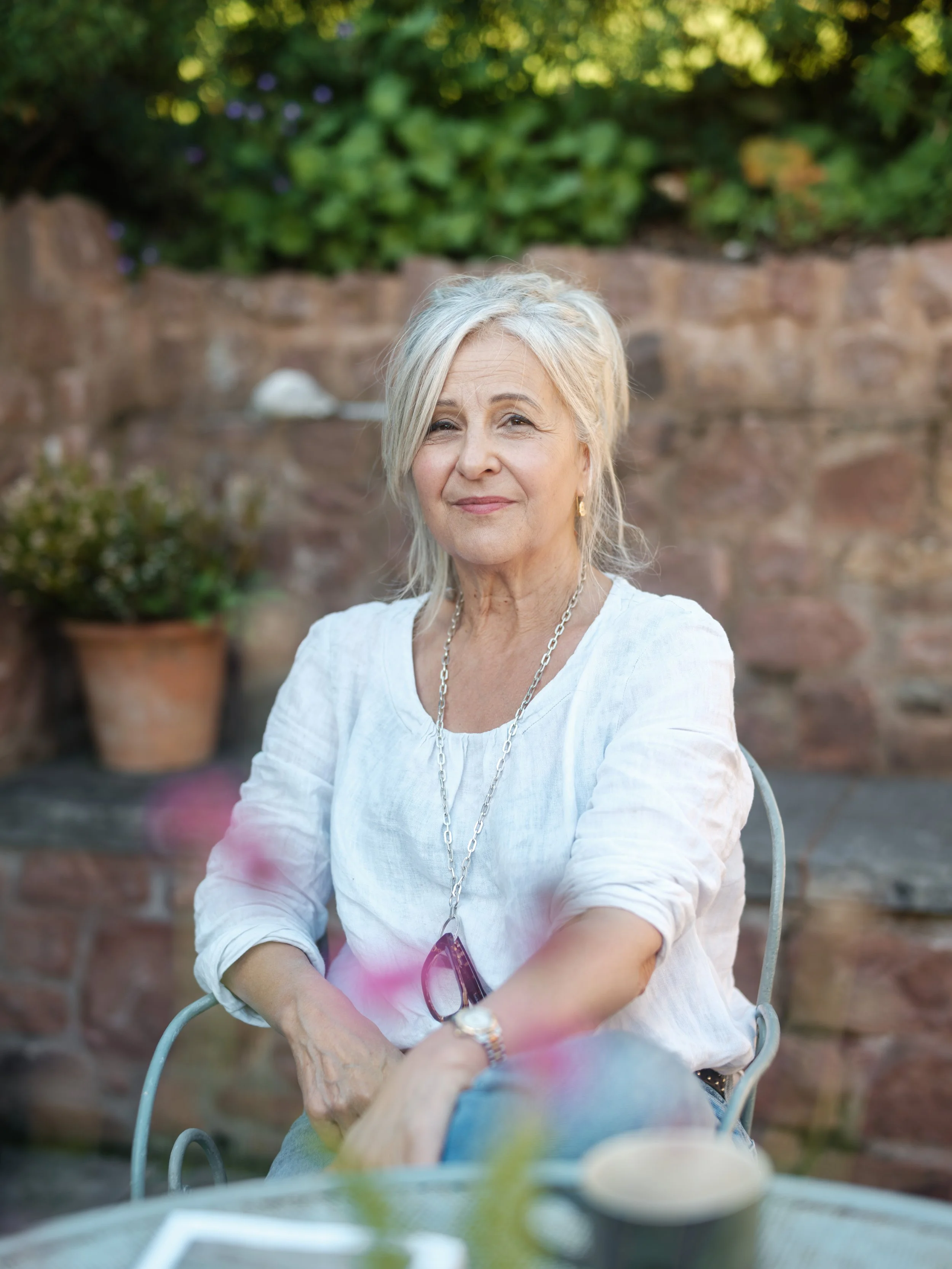 An older woman with blonde hair sitting outdoors at a table, wearing a white blouse, and smiling softly.