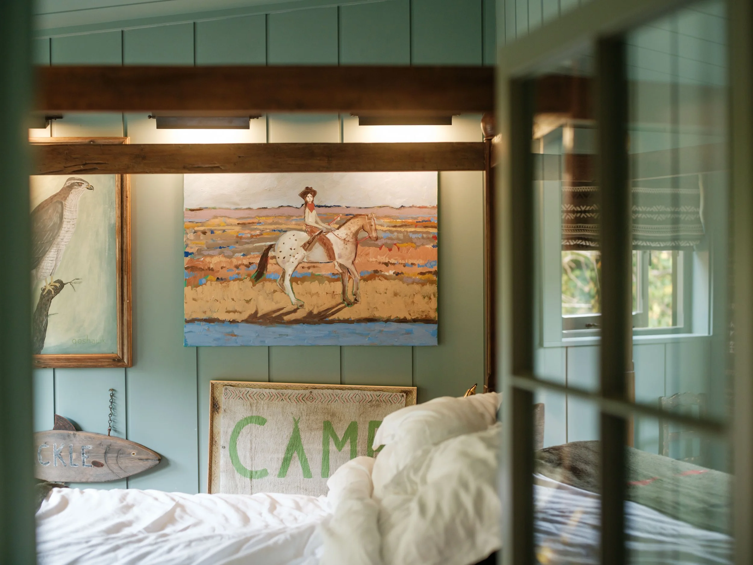 View of a cozy bedroom with paintings on the wall, including a girl riding a horse, a bird illustration, and a sign that says 'CAMP'. Part of a bed with white bedding is visible in the foreground, with a window showing greenery outside.