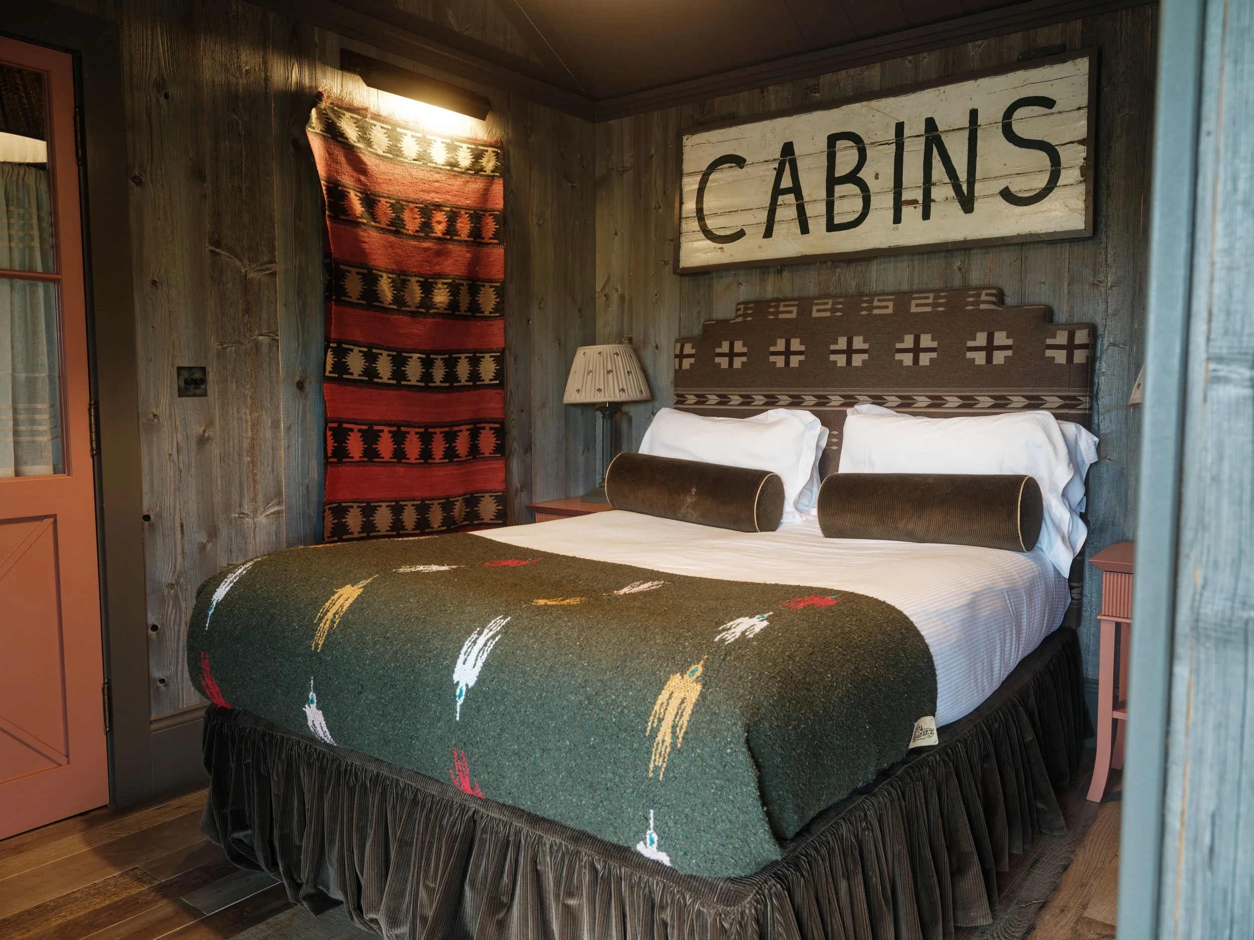 A cozy bedroom with a bed featuring a white sheet, brown pillows, and a patterned blanket with leaf motifs. The room has rustic wood paneling, a red and black decorative wall hanging, a lamp on the nightstand, and a large sign above the bed that says