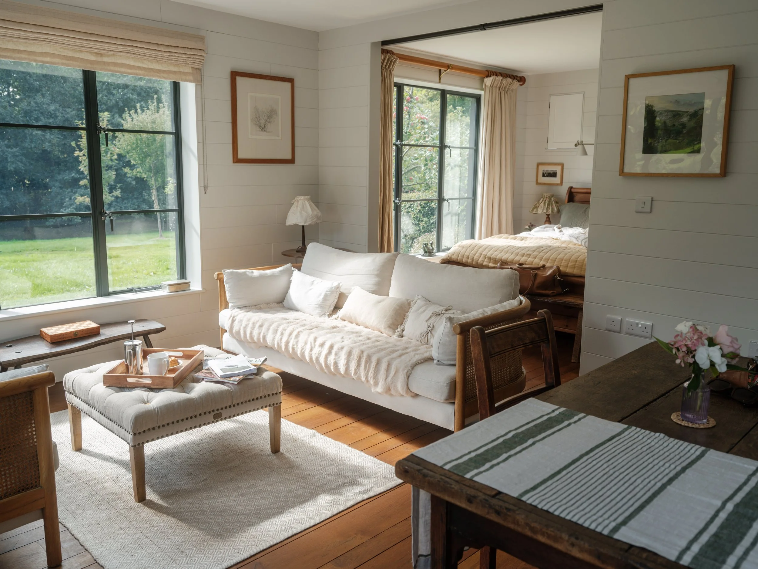 Bright living room with a white sofa, a wooden coffee table with a tray, a beige rug, and large windows with curtains overlooking a green lawn and trees. In the background, there is a bedroom with a bed, framed artwork, and a window.