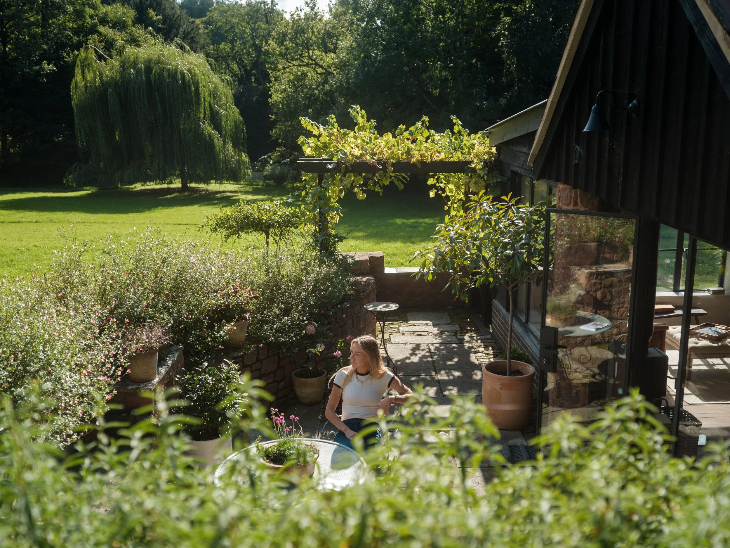 A young woman sitting at a patio table outside a house, surrounded by potted plants and greenery on a sunny day.