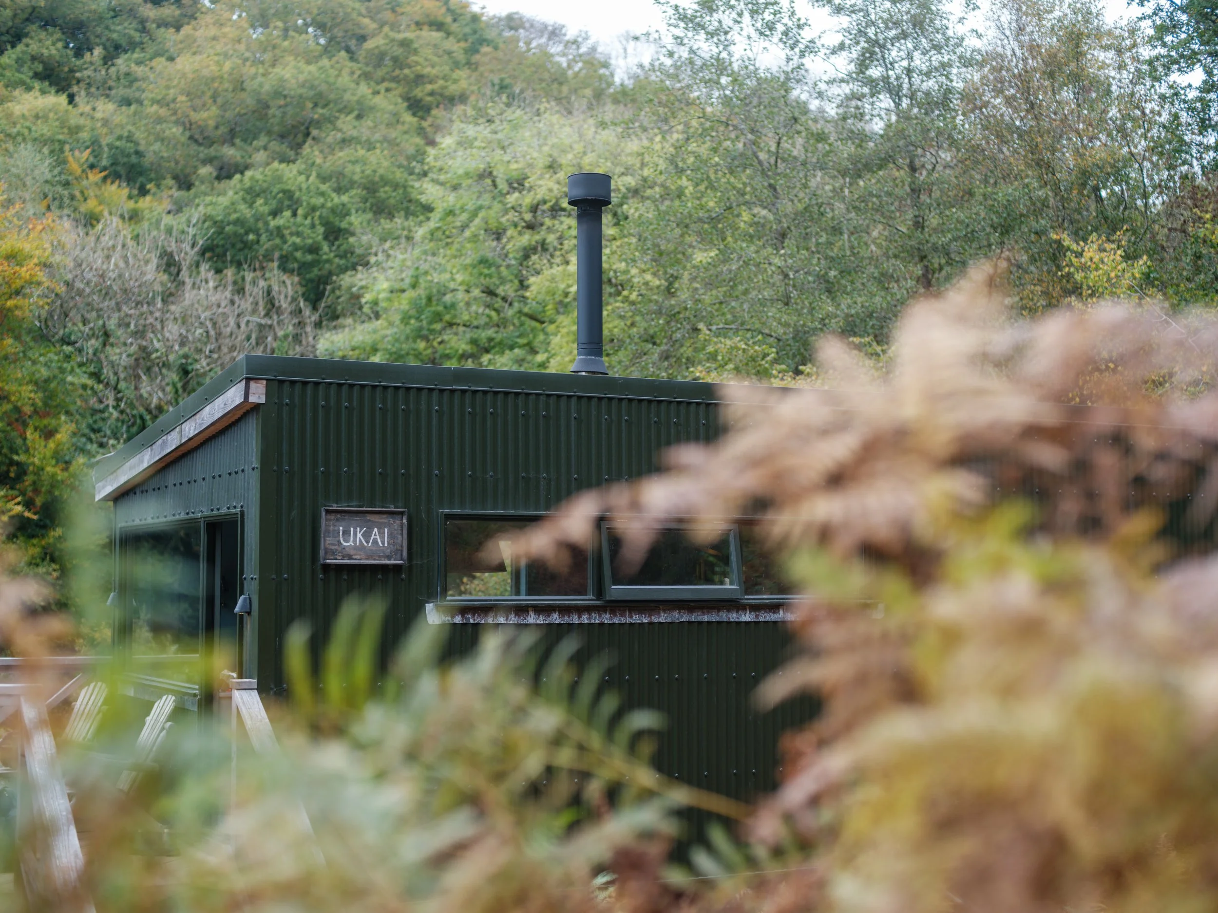 A black building with a chimney, labeled 'UKAI', is set against a backdrop of green trees and hills, with some foreground foliage partially obscuring the view.