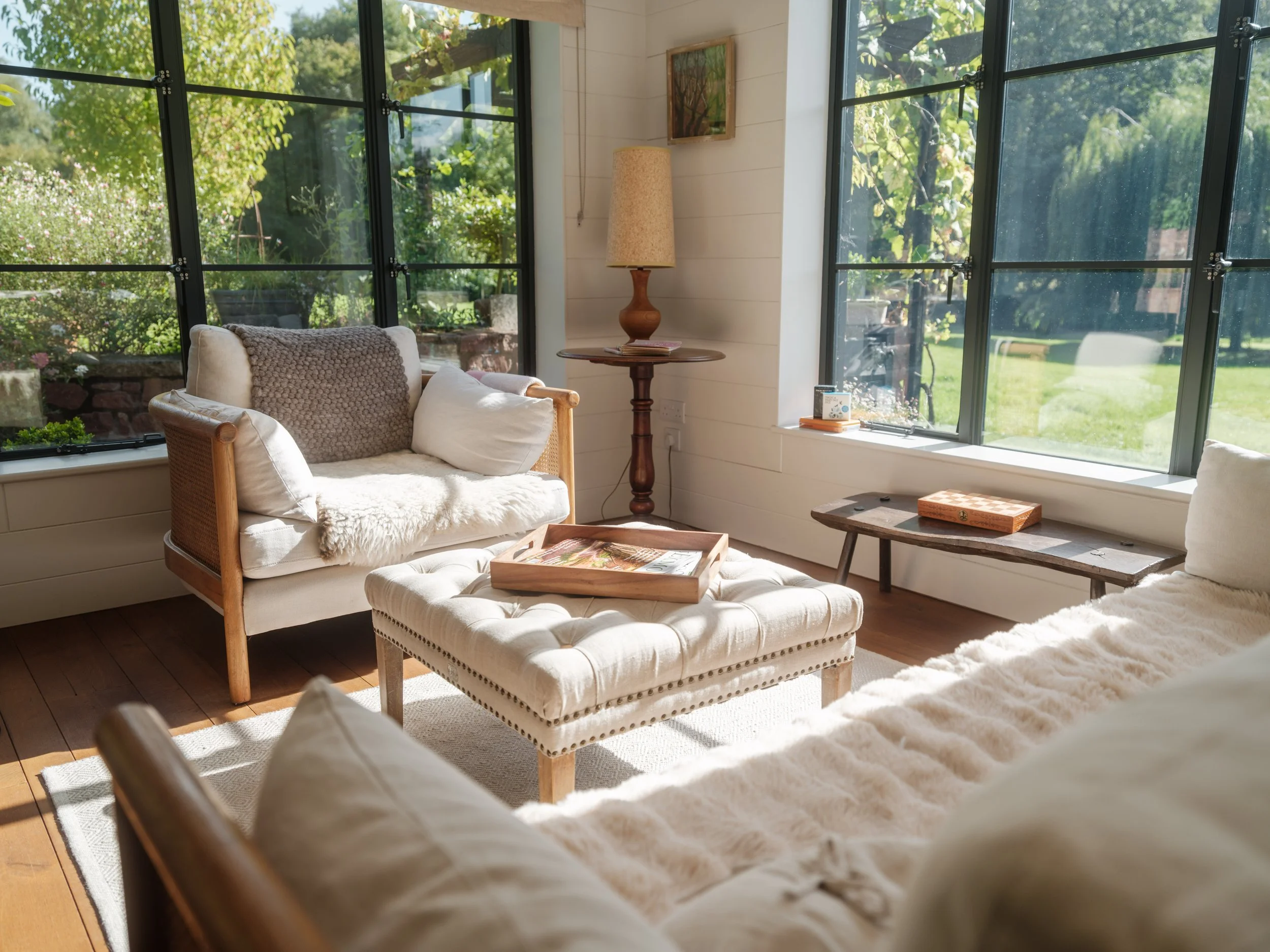 Cozy living room with large windows overlooking green garden, beige sofa with white and brown pillows, a tufted ottoman, wooden side table with a lamp, and natural sunlight filling the space.