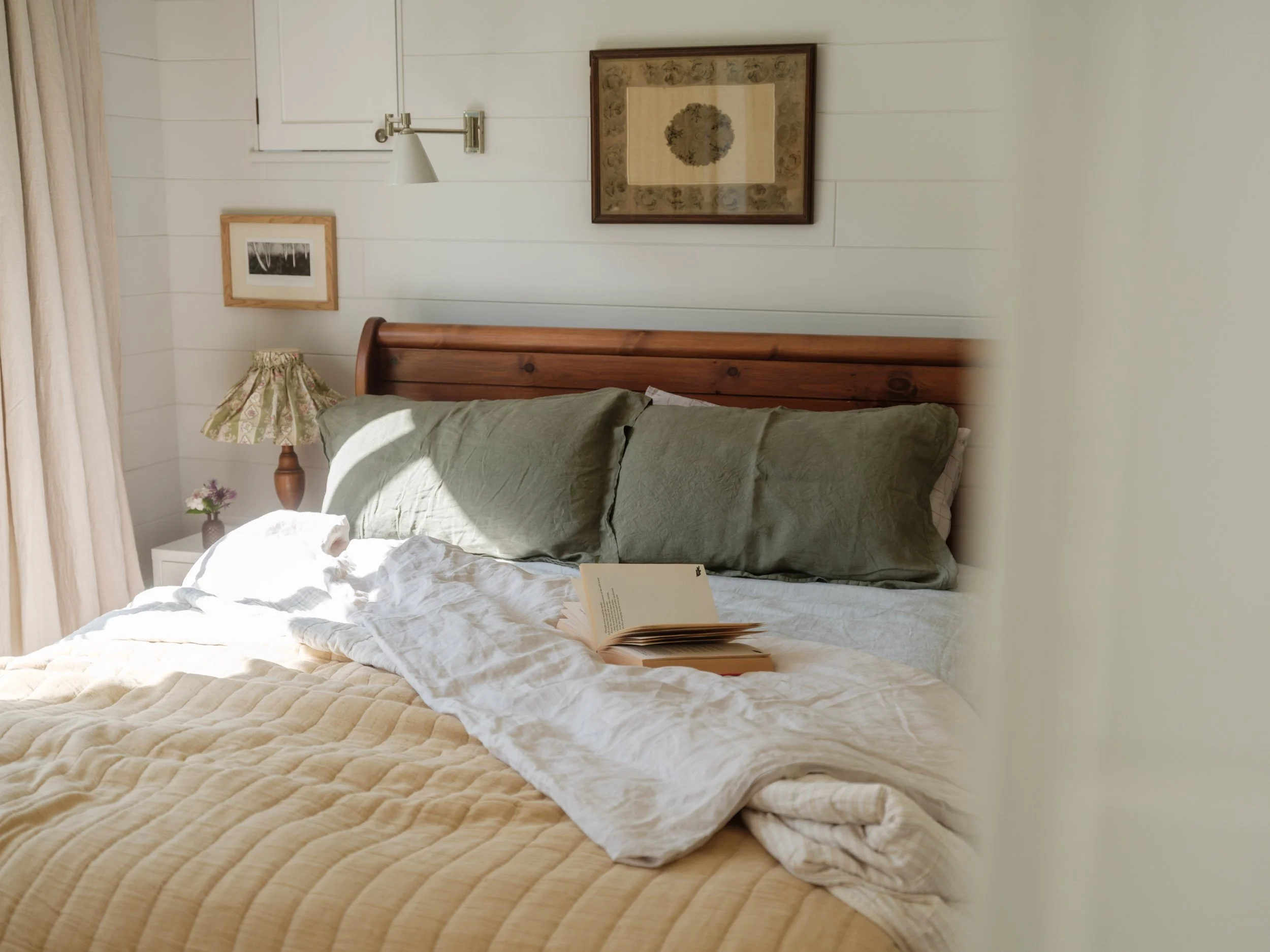 Cozy bedroom with a wooden bed frame, green pillows, white sheets, a book on the bed, a nightstand with a small flower vase, a floral lampshade, wall art, and curtains, illuminated by natural sunlight.