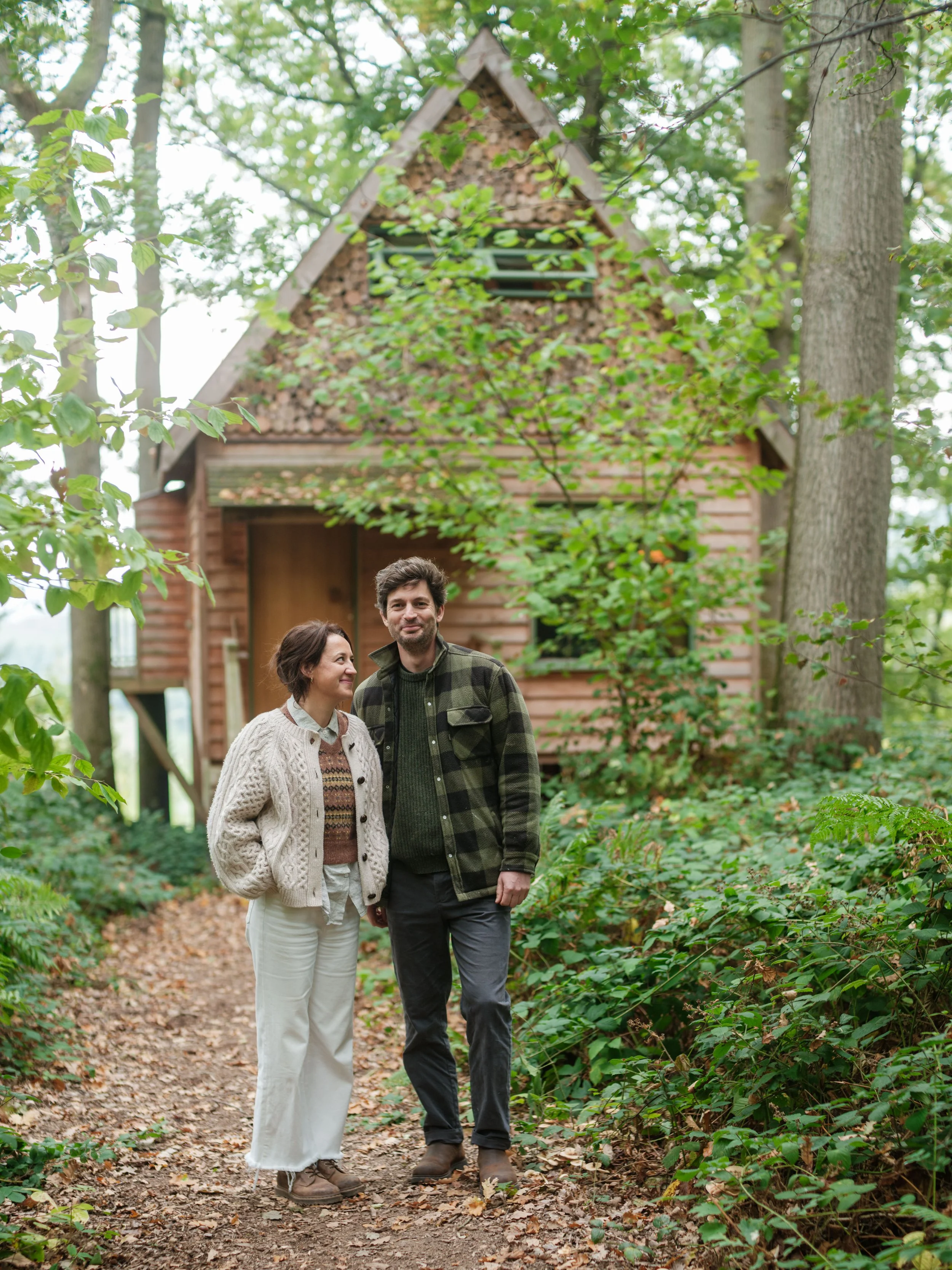 A couple standing on a wooded trail in front of a small log cabin, surrounded by green foliage.