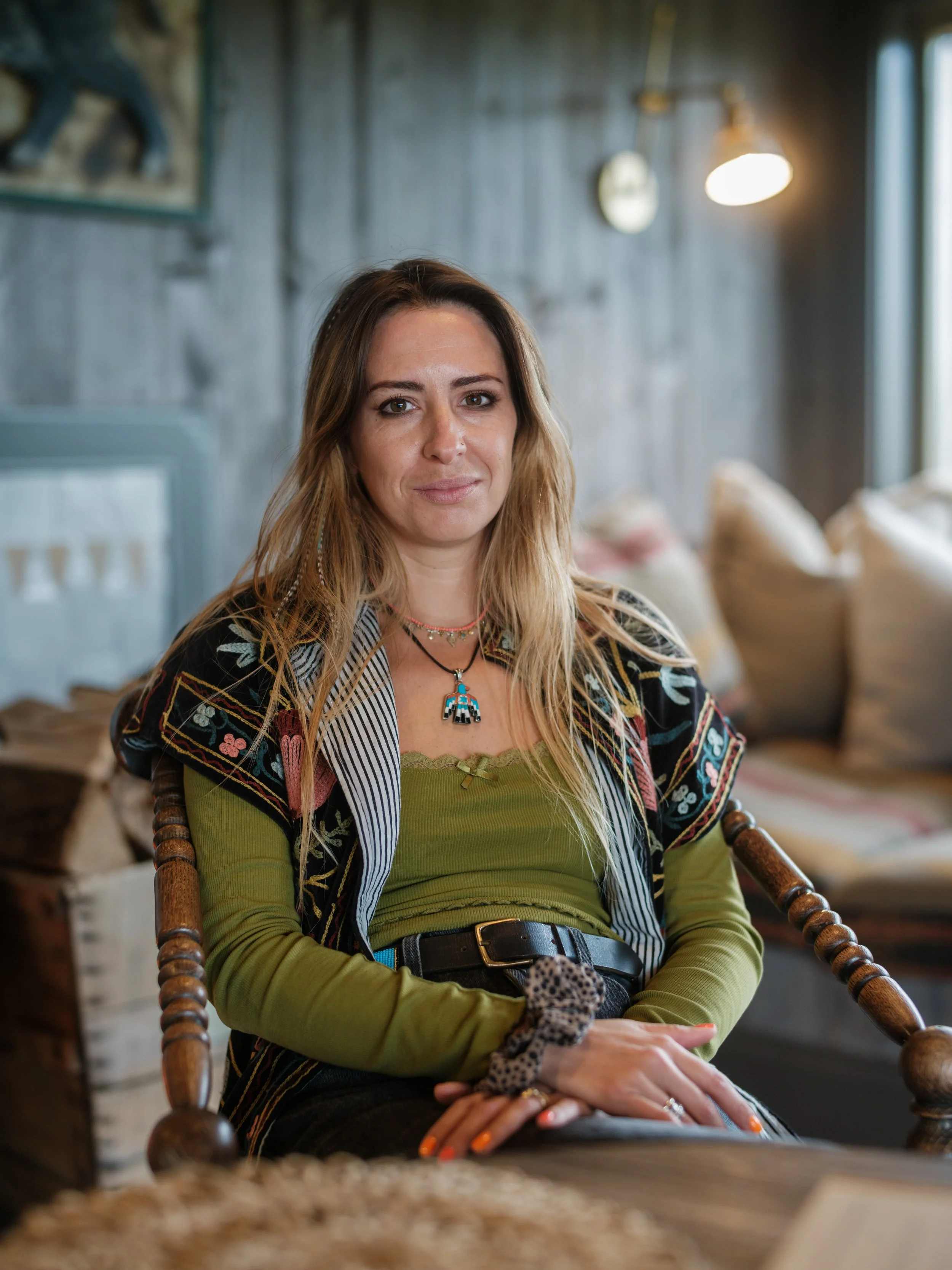 A woman with long, light brown hair sitting in a wooden chair in a cozy, rustic room with wooden walls and soft lighting, wearing a green top, layered necklaces, and a leopard print scrunchie.