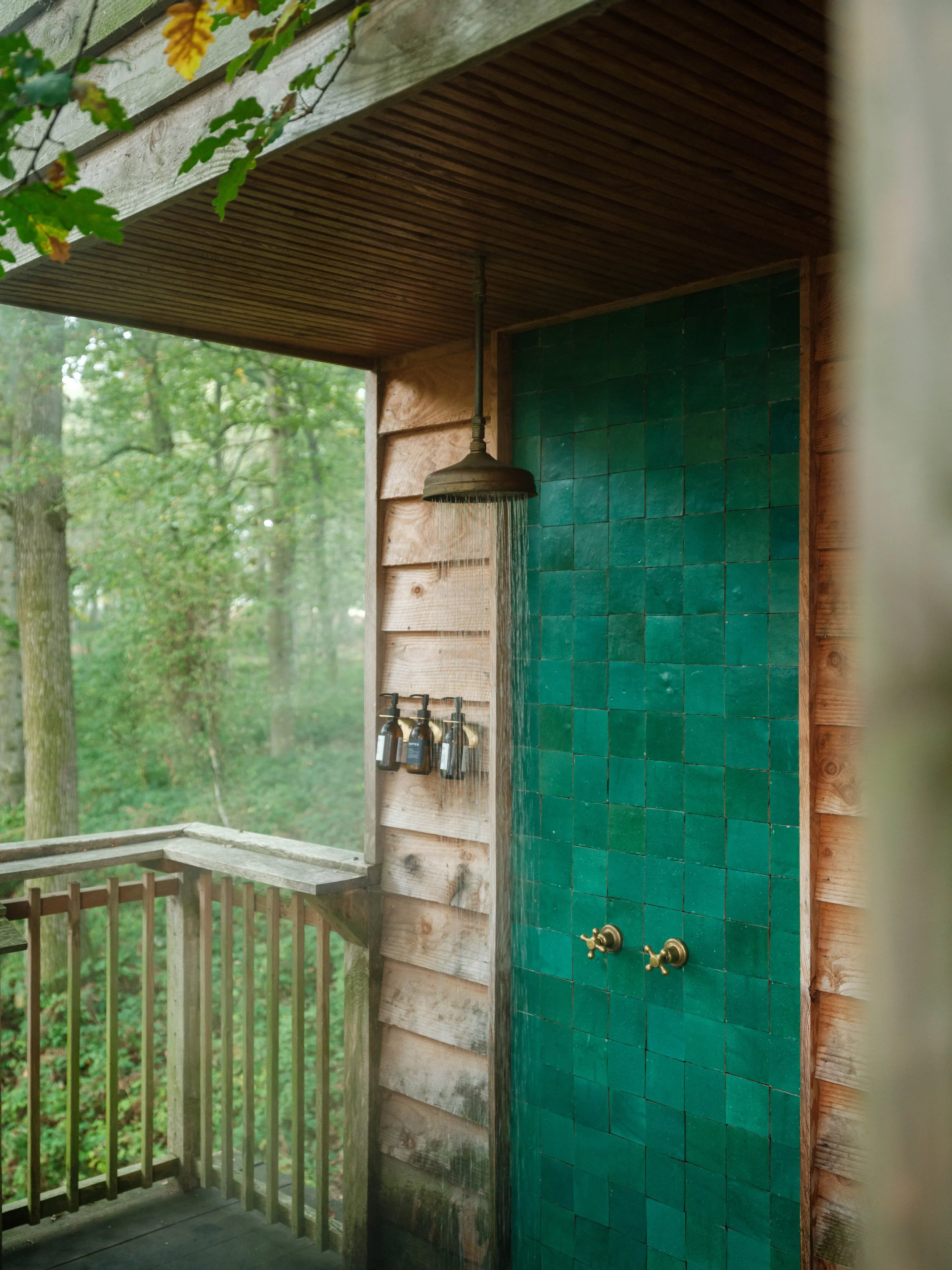 Outdoor shower with two brass taps on a green tiled wall, surrounded by a wooden deck and railing, in a wooded area.