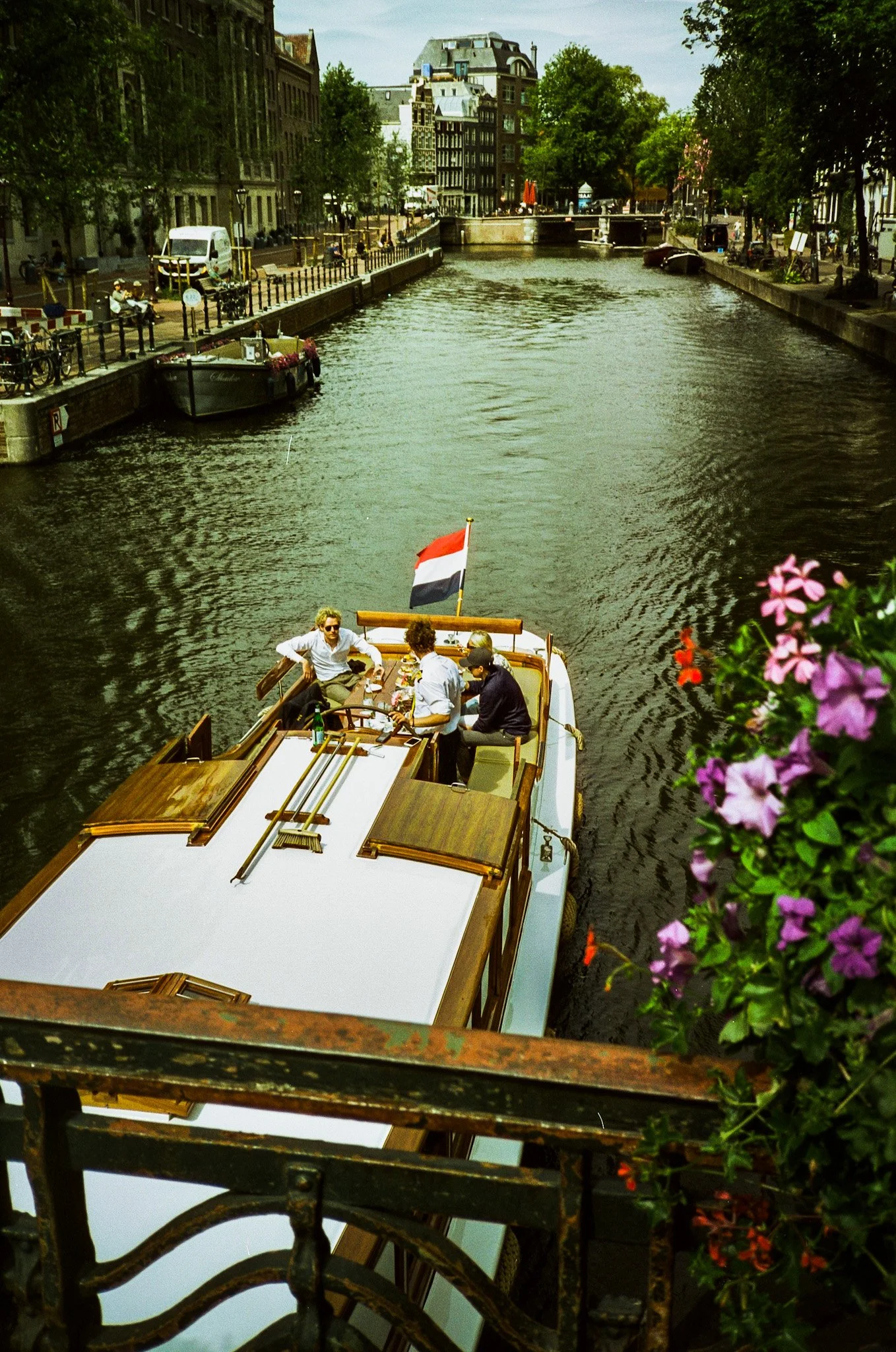 People on a boat with a red, white, and black flag on a canal in an urban area with buildings, trees, and flowers.