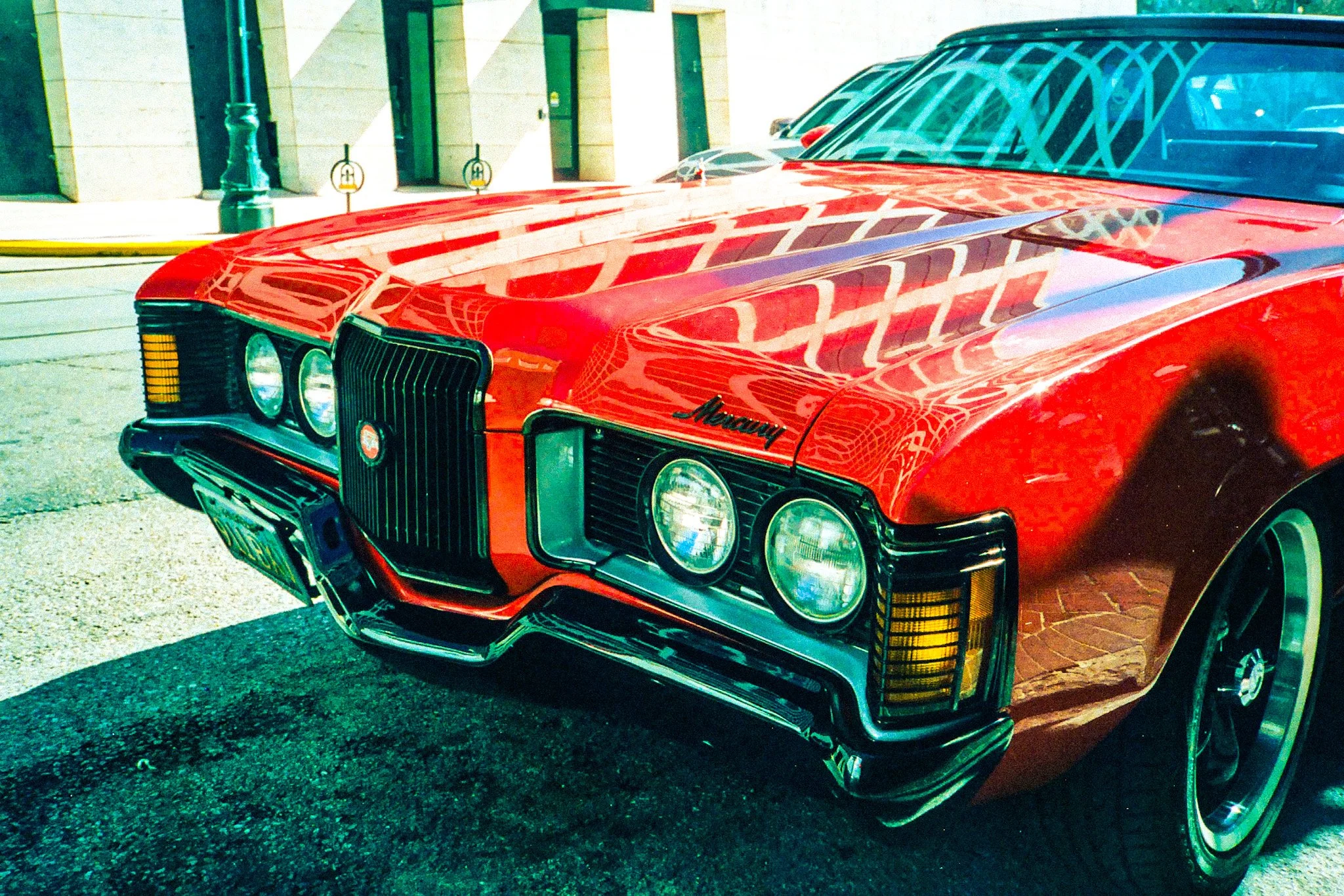 Close-up of a vintage red Mercury car parked outside buildings, showing the front and part of the side with reflections of nearby structures on its shiny surface.