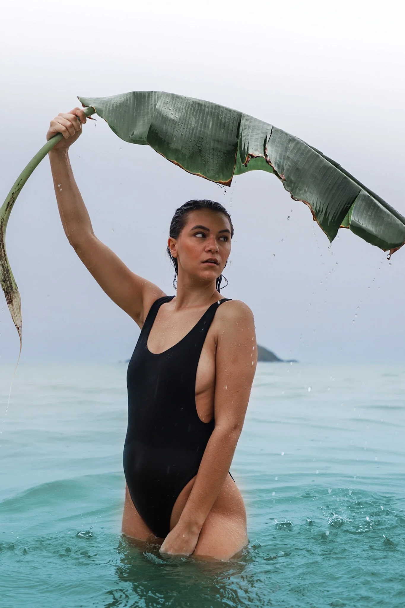 A woman in a black swimsuit stands in the ocean holding a large banana leaf above her head.