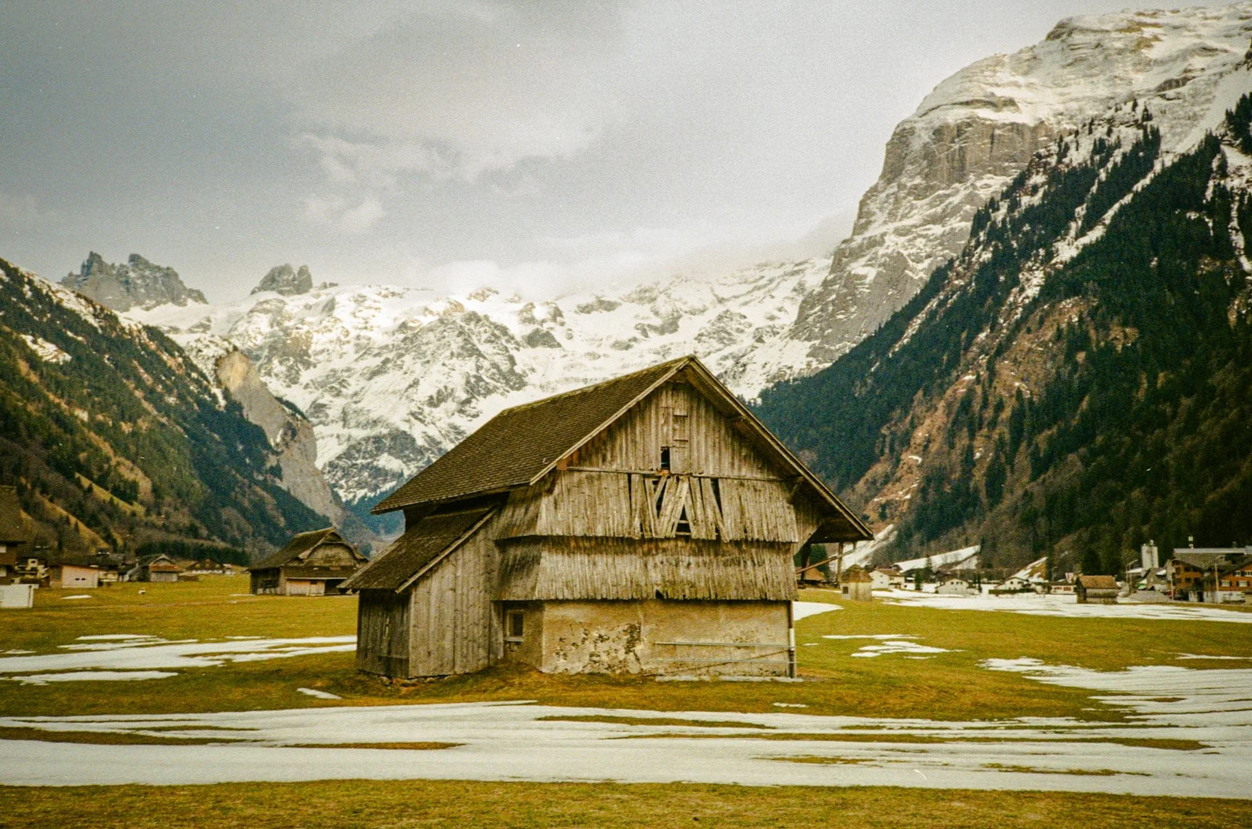 An old wooden barn in a valley with snow patches on the grass, surrounded by mountains with snow and trees.