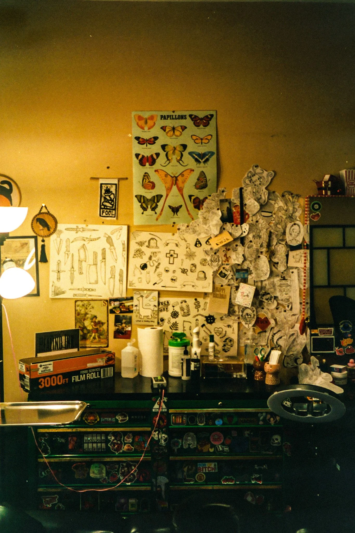 A room with a wall decorated with butterfly posters and sketches, a black table with various items, and a pinball machine with stickers.