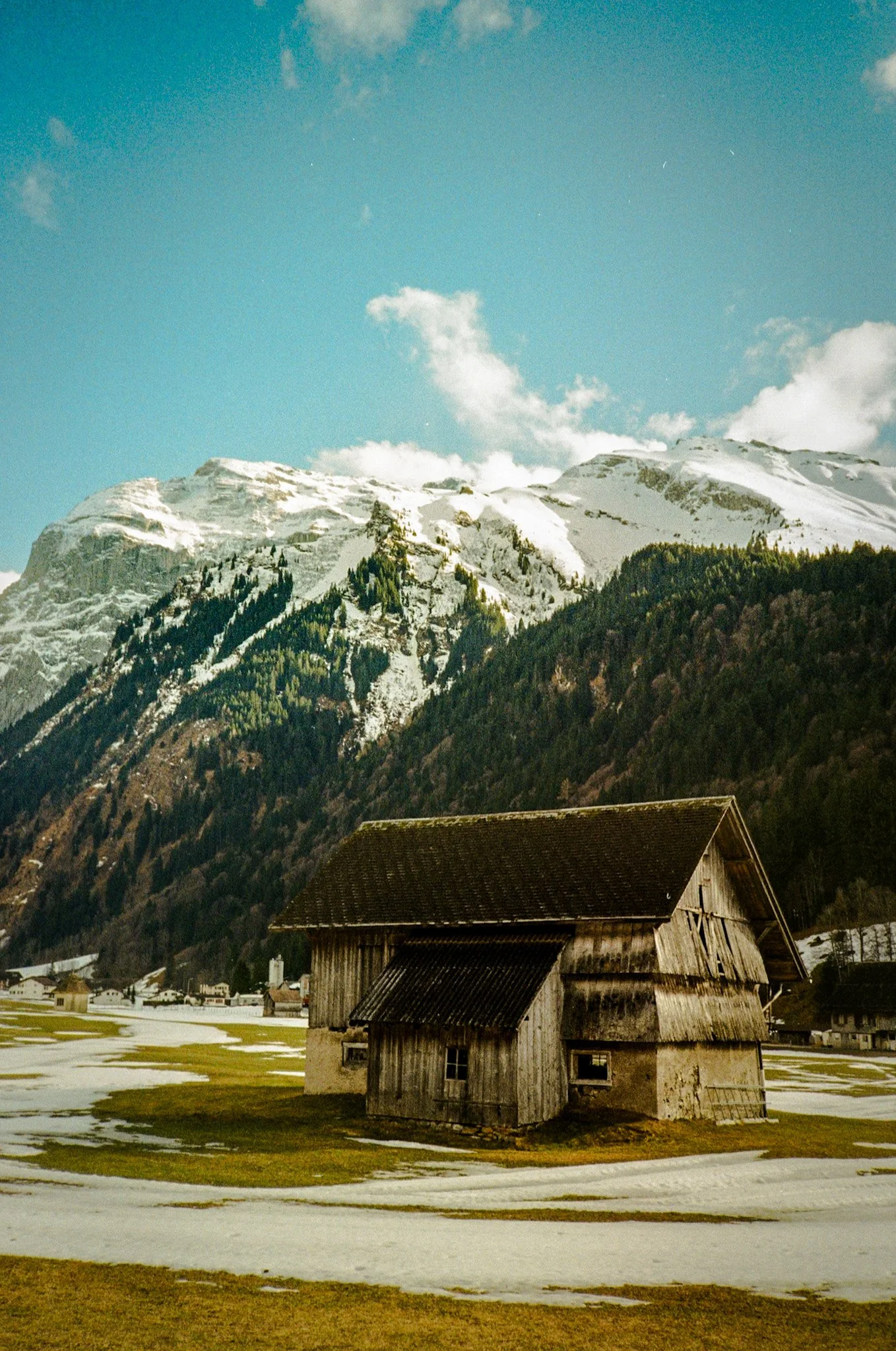 A rustic wooden barn in a snowy field with snow-capped mountains and a partly cloudy blue sky in the background.