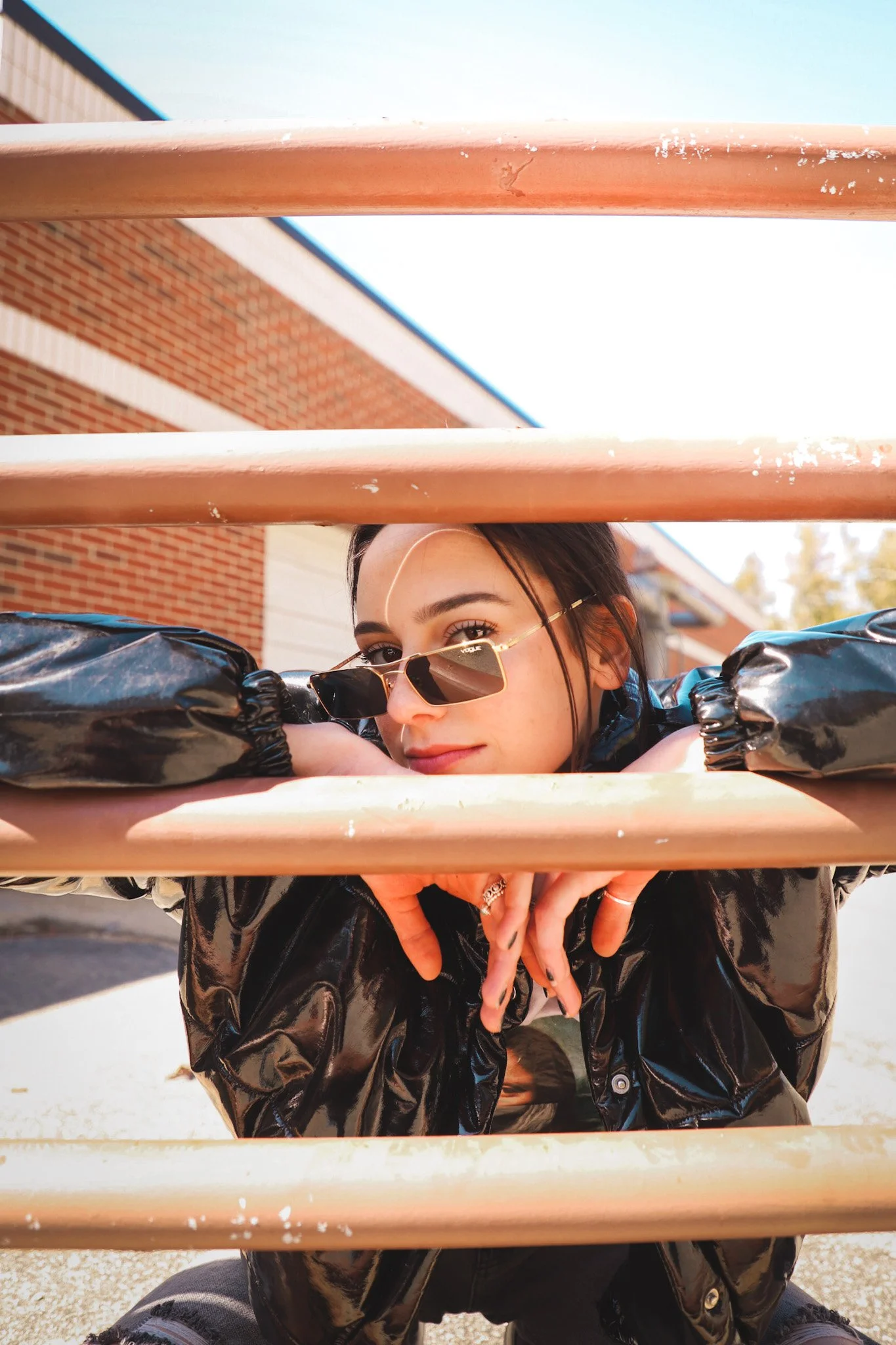 A young woman with dark hair wearing sunglasses and a black shiny jacket, resting her head and arms on a peach-colored metal railing, with a brick building and blue sky in the background.