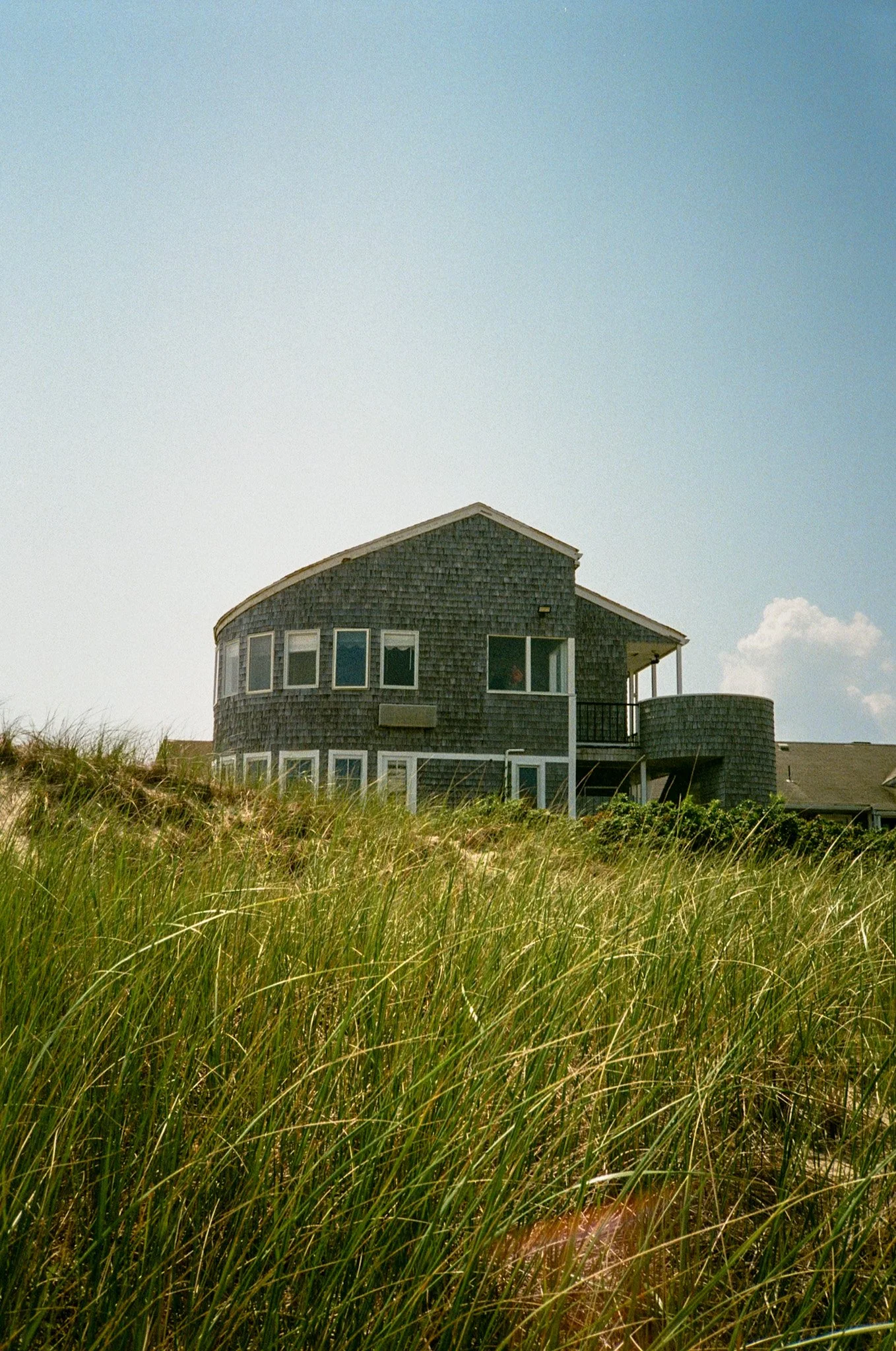 A house with dark wooden shingles sits on a grassy dune under a blue sky with some clouds.