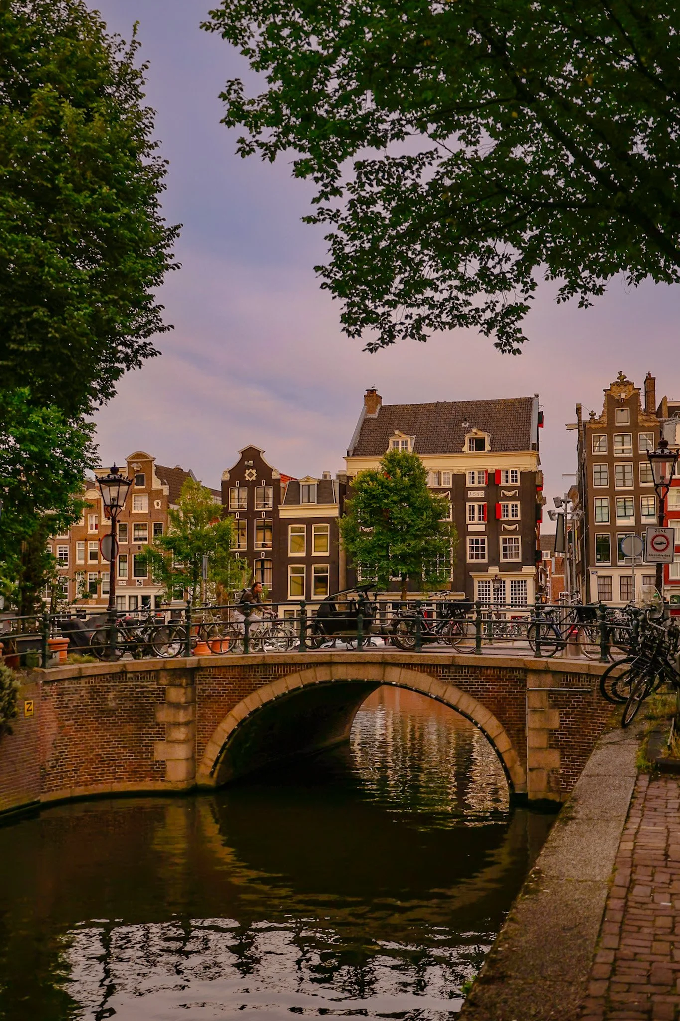 A canal with a small arched bridge over it, lined with bicycles and trees, with historic buildings in the background and an overcast sky.