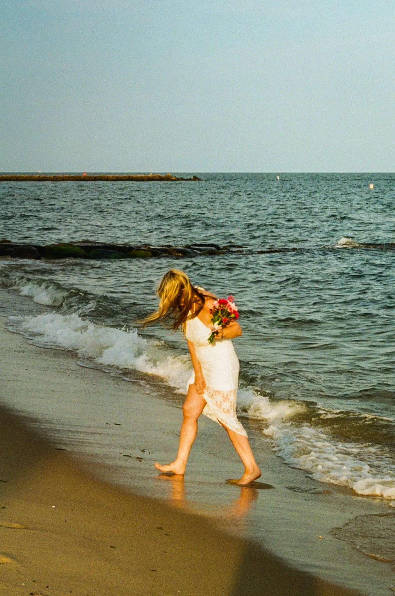 A woman in a white lace dress holding a bouquet of pink flowers walking barefoot on the beach.