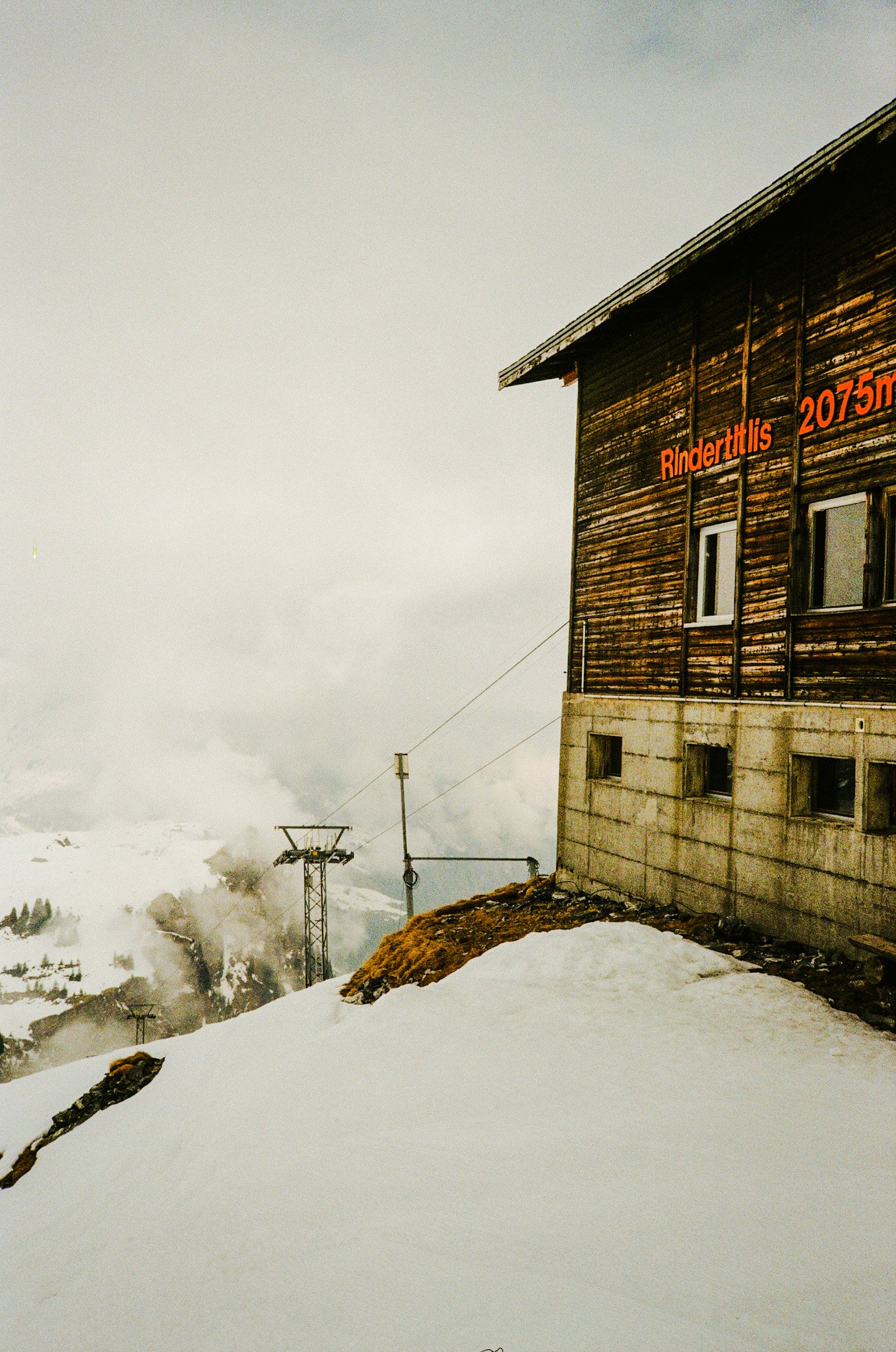 A mountain hut with wooden siding and small windows, snow covering the ground, ski lift towers, and cloudy sky in the background.