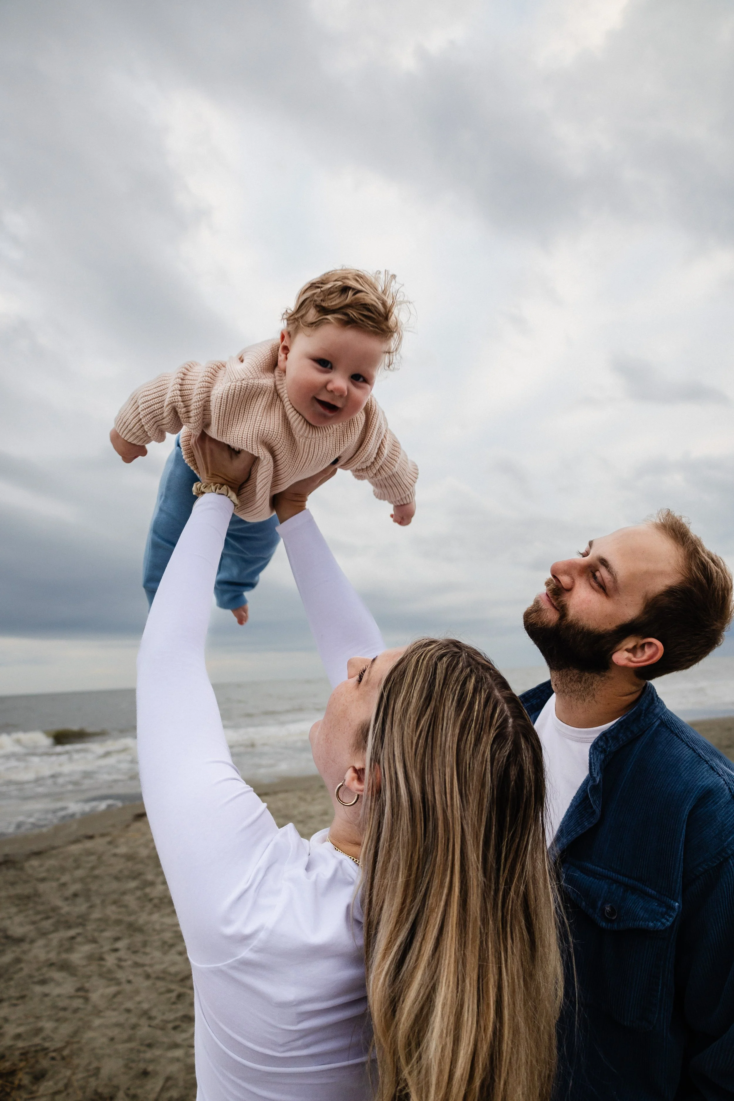 A woman lifts a young boy in the air on a beach, with a man watching and smiling under a cloudy sky.