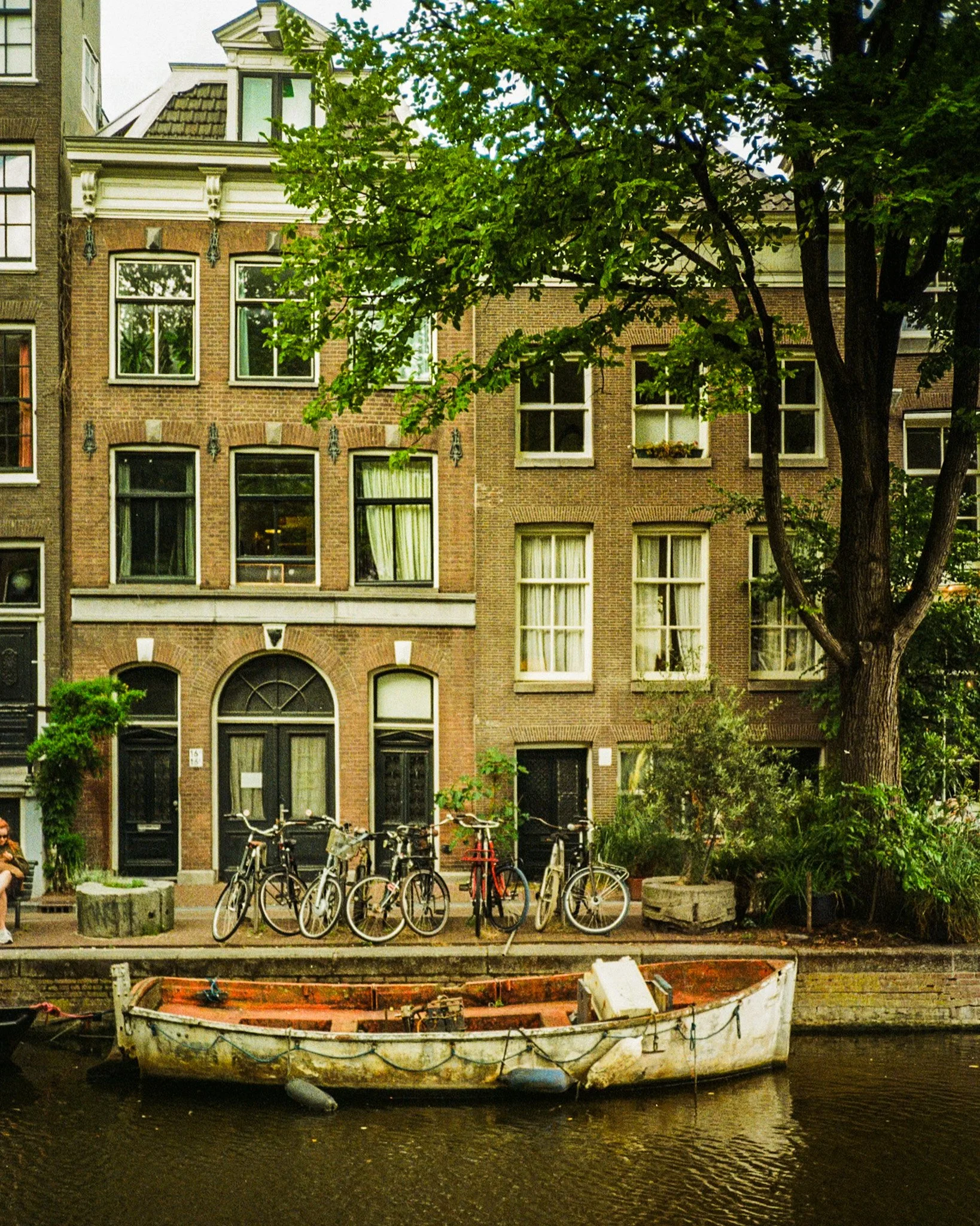 Bicycles parked along a canal with an old boat in the water, brick buildings, and a tree in an urban setting.