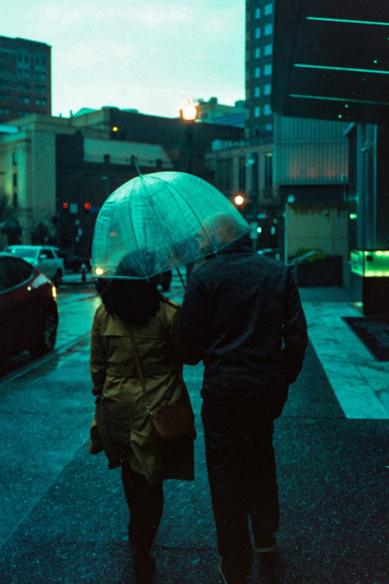 A couple walking under a transparent umbrella on a rainy city street at dusk.