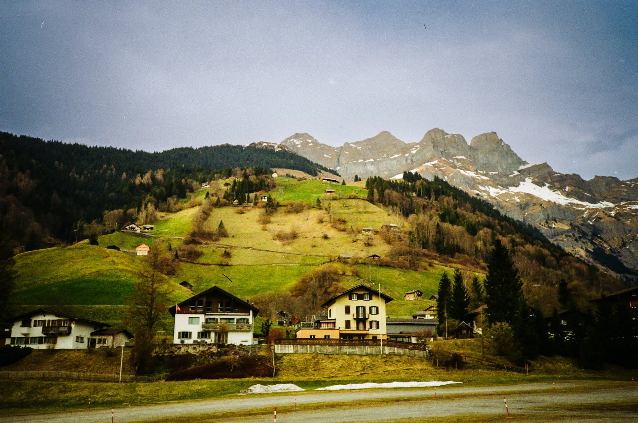 Mountains with snow patches, green hillside with scattered houses, and a cluster of houses in the foreground.